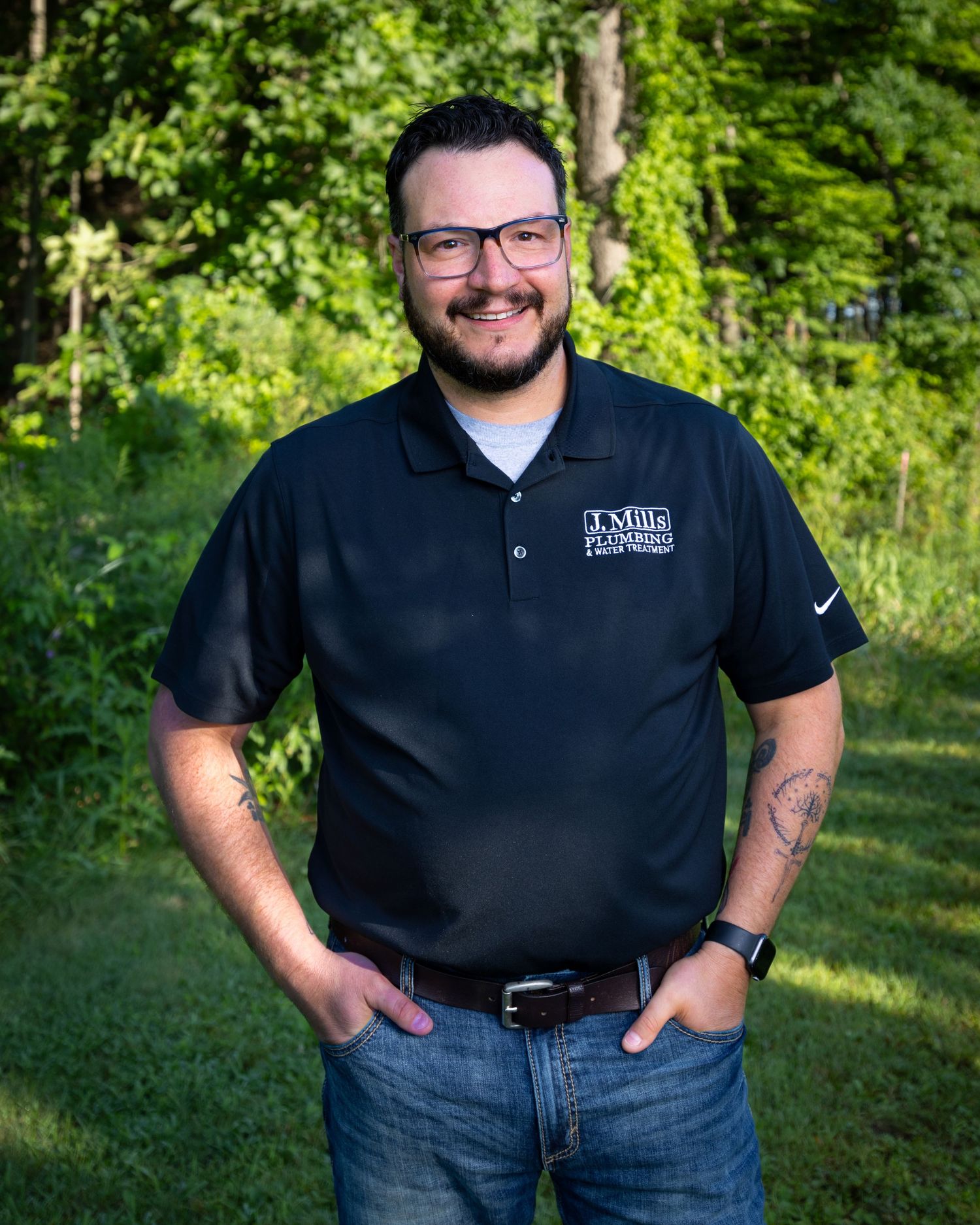 Professional outdoor portrait in black polo shirt and jeans against green foliage.