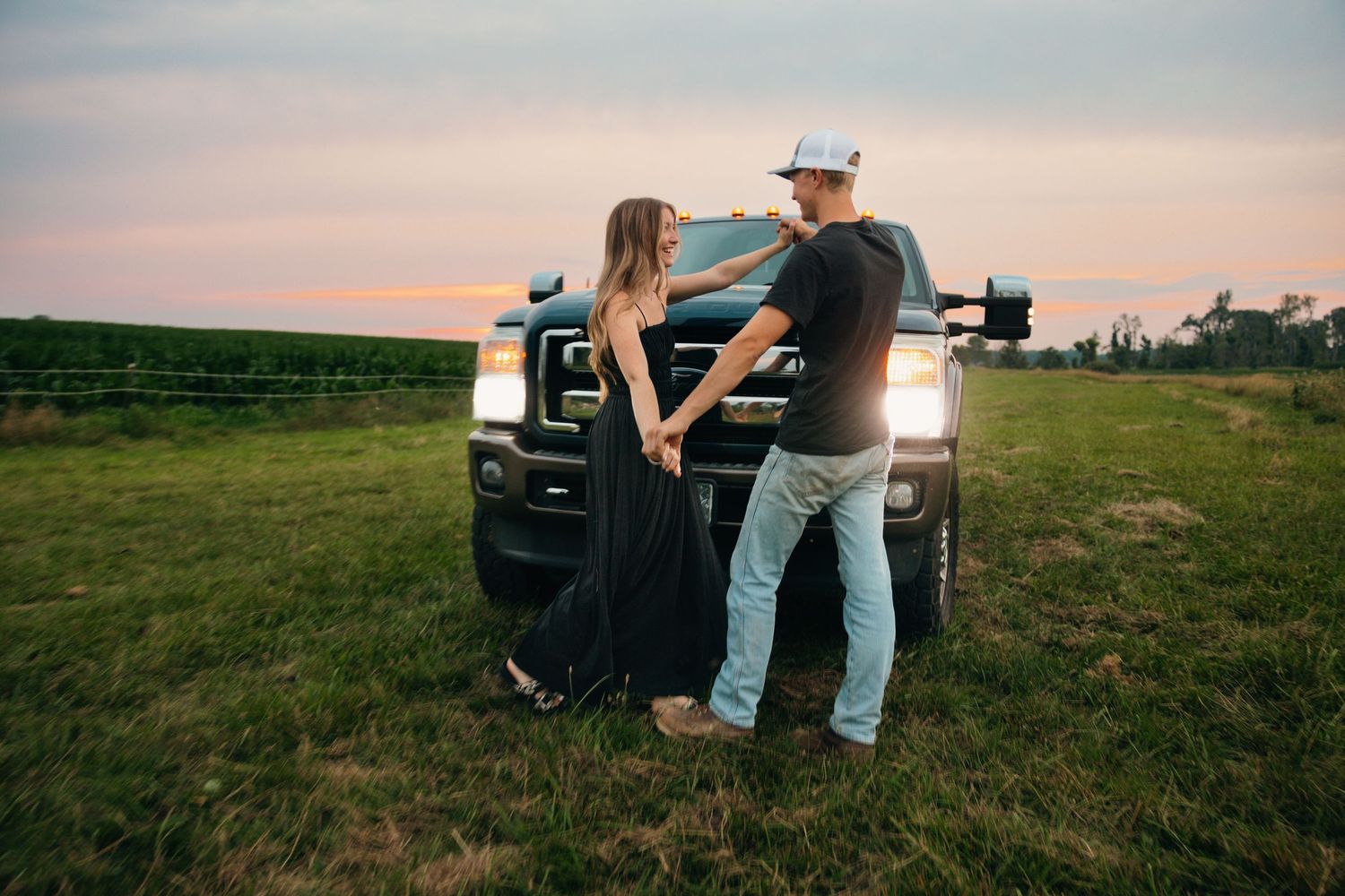 Couple standing together in front of a black pickup truck at sunset in a grassy field.