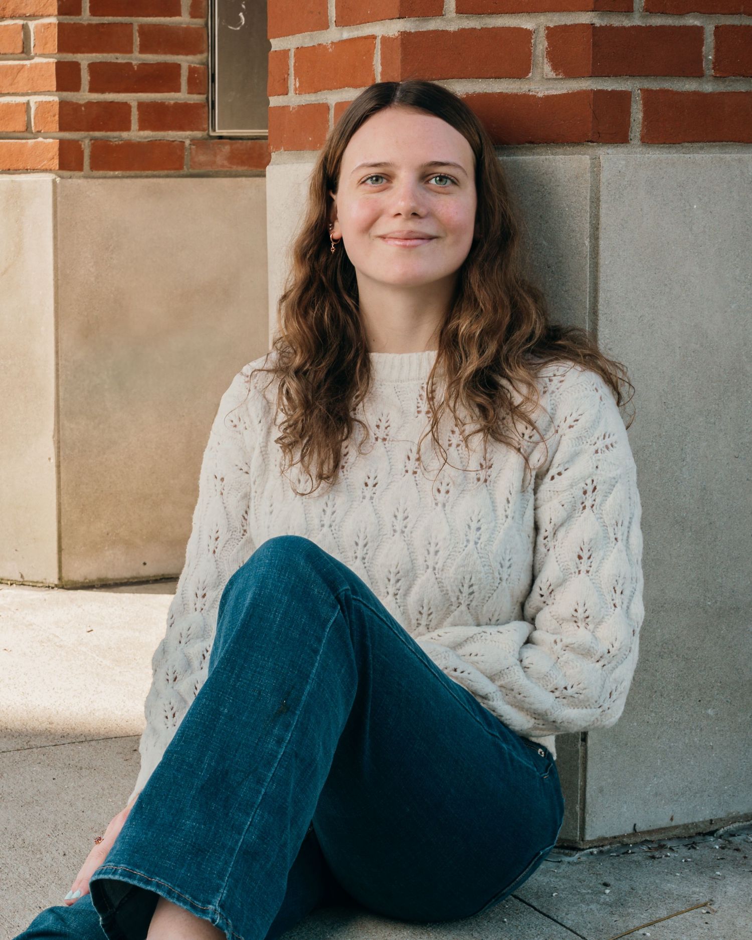 Portrait sitting by brick wall wearing white knit sweater and blue jeans.