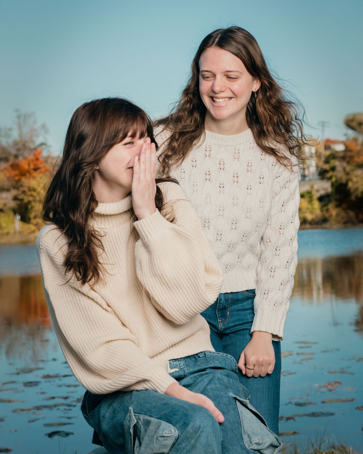Two friends sharing a joyful moment by a lake during fall with cozy sweaters.