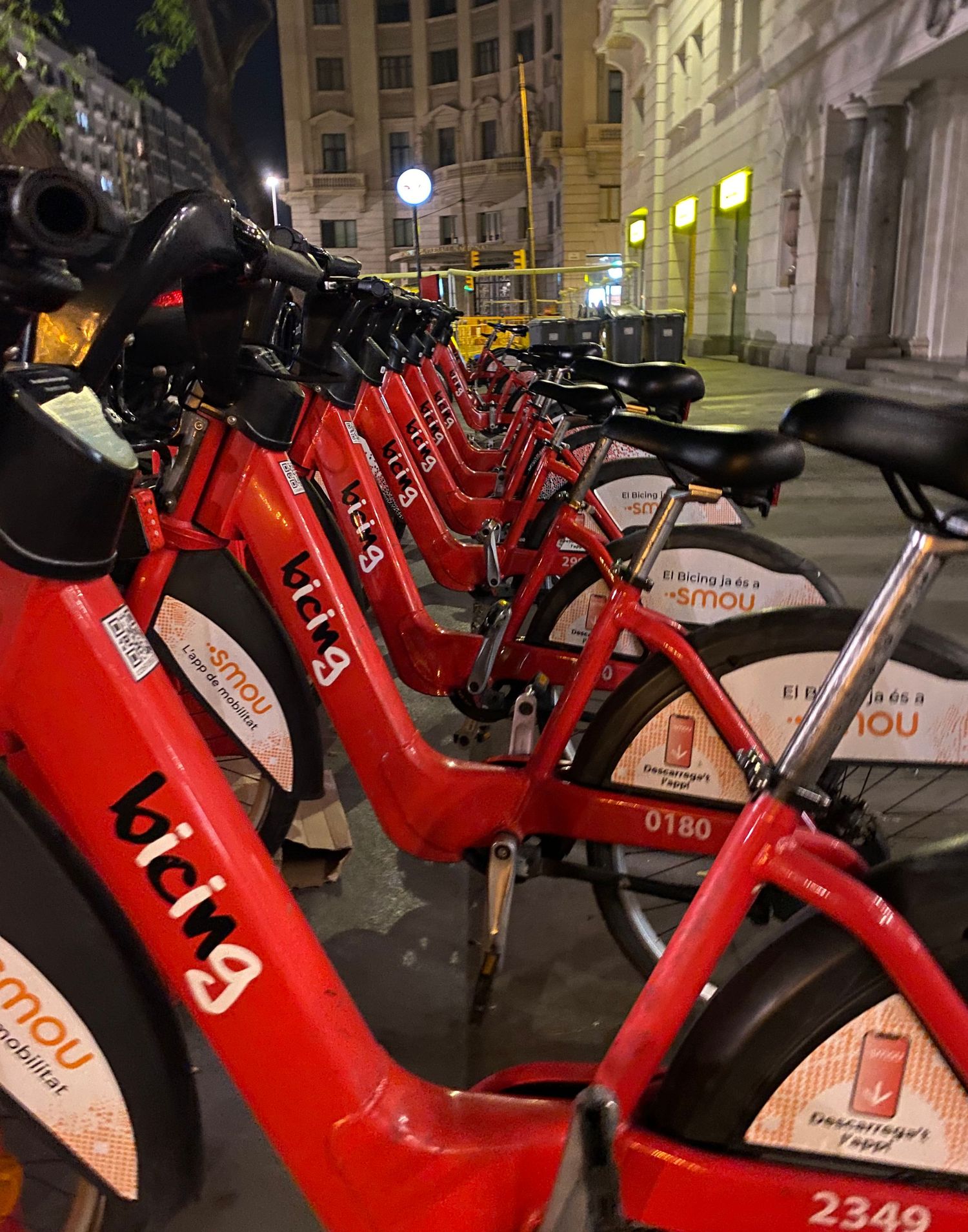 A row of red rental bikes parked in a docking station at night with city buildings in the background.