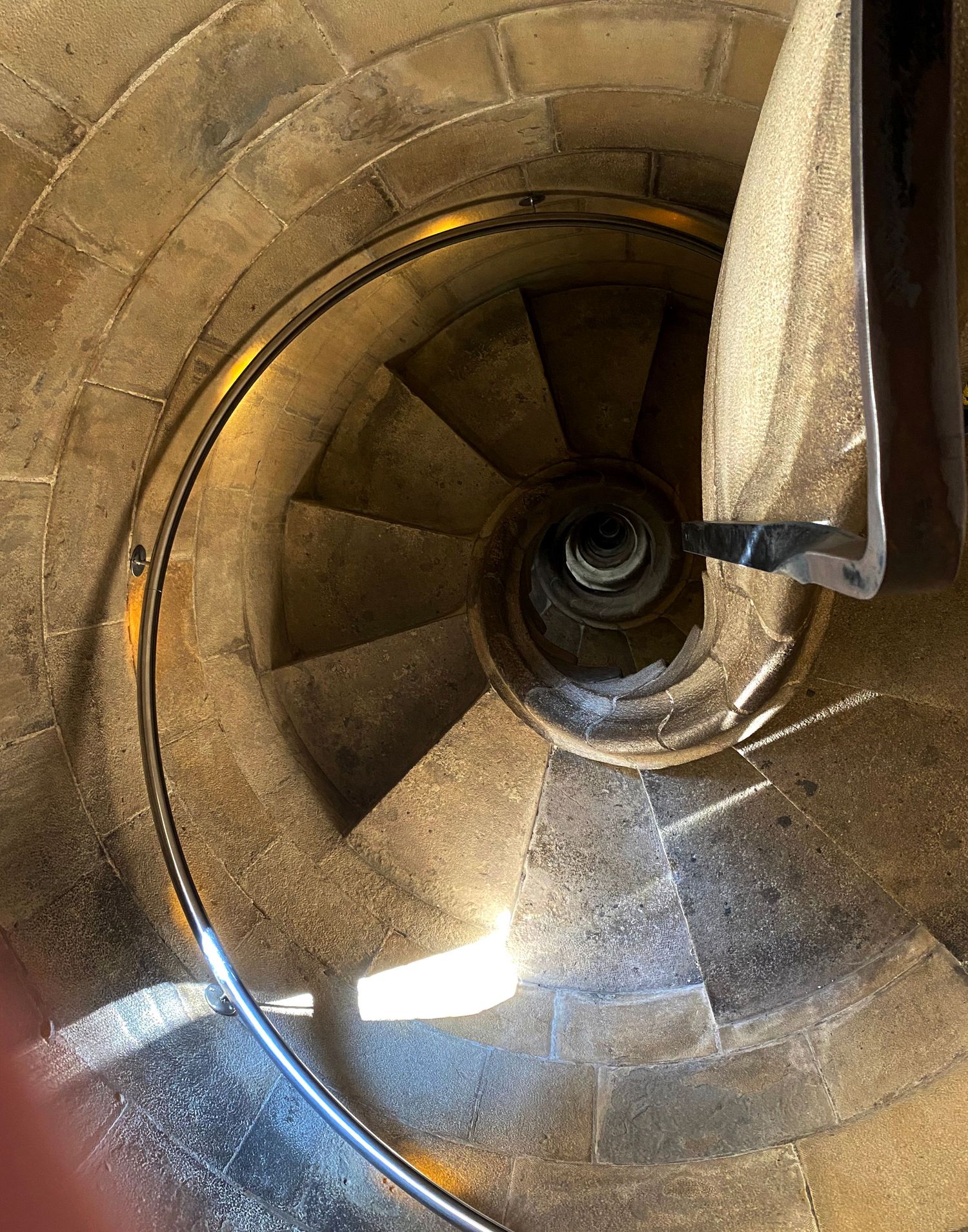 A spiral stone staircase descends in a circular pattern with sunlight filtering through from above.