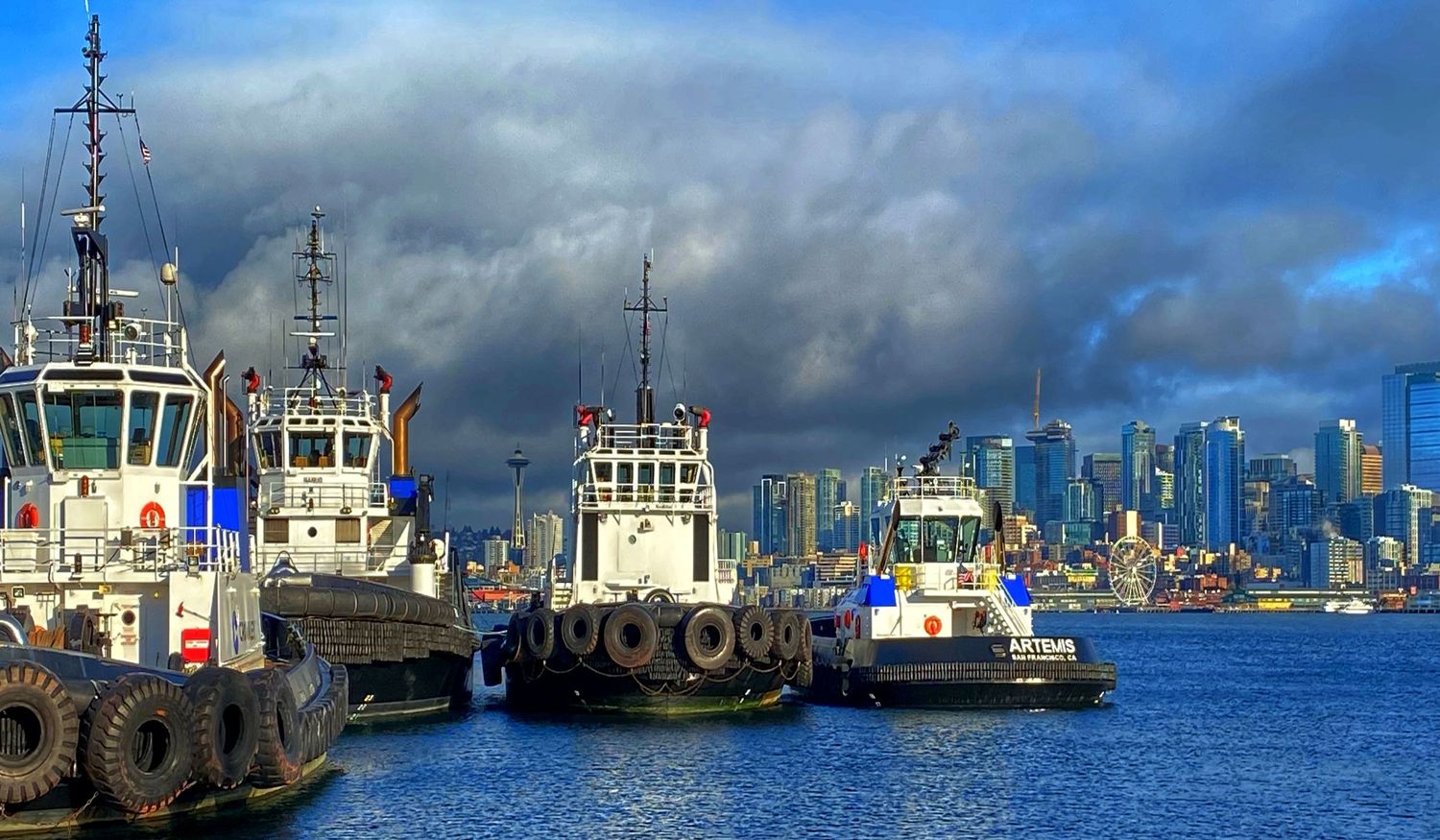 Panoramic view of boats docked in a harbor with city skyline and dramatic clouds.