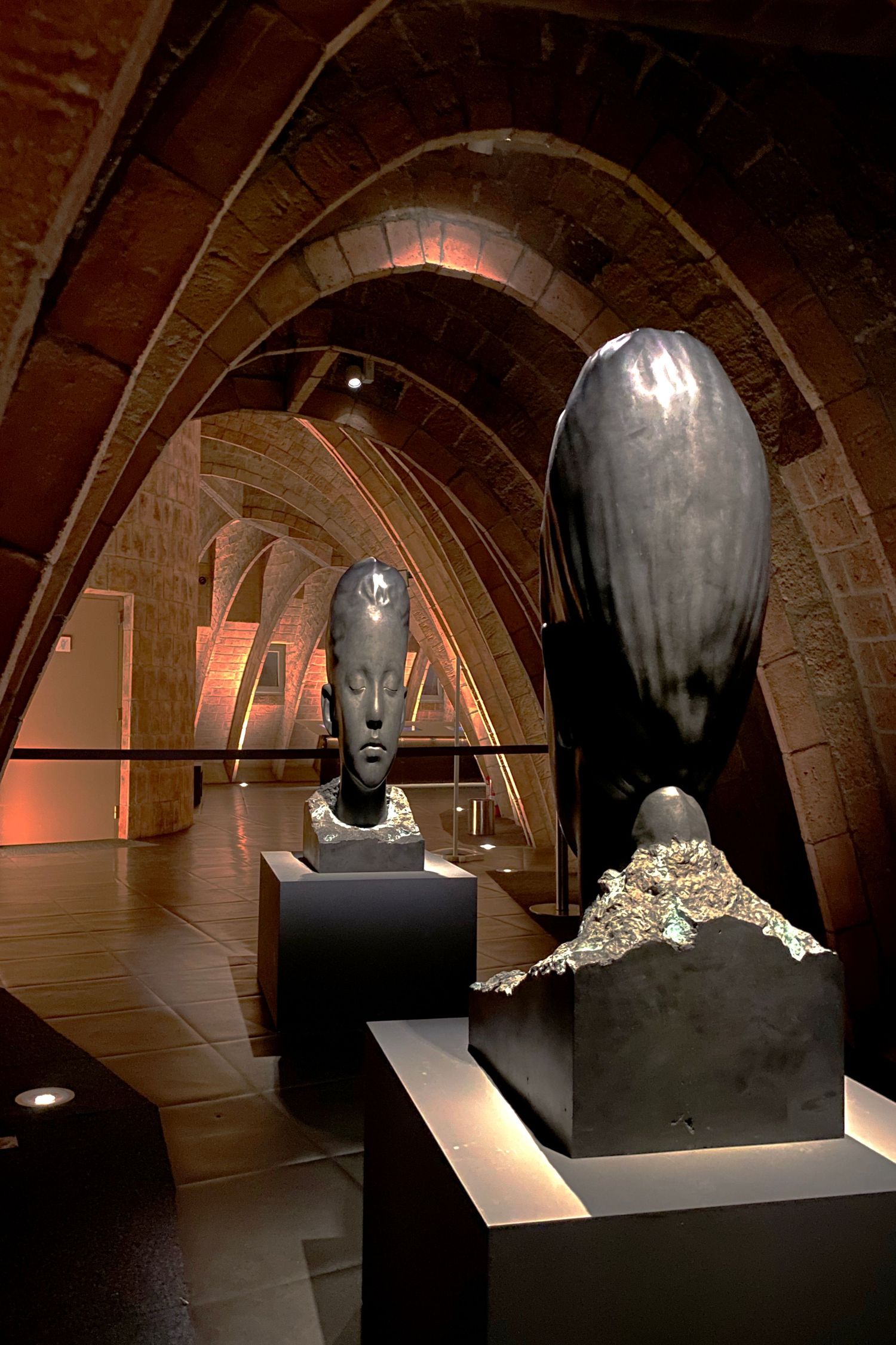 Dark vaulted gallery space displaying sculptural busts on pedestals with dramatic lighting.