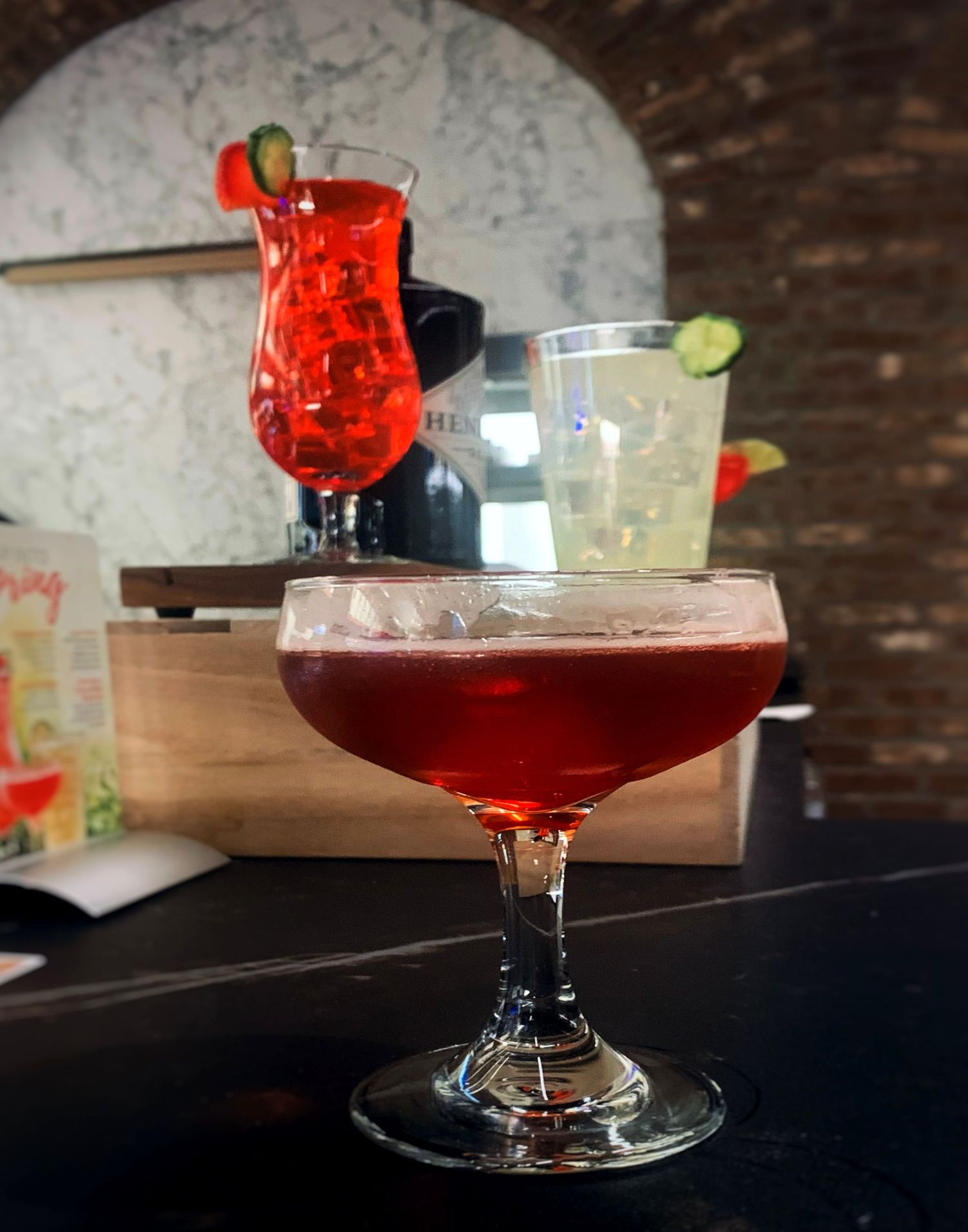 Red cocktail in coupe glass garnished with fruit on dark wooden surface.