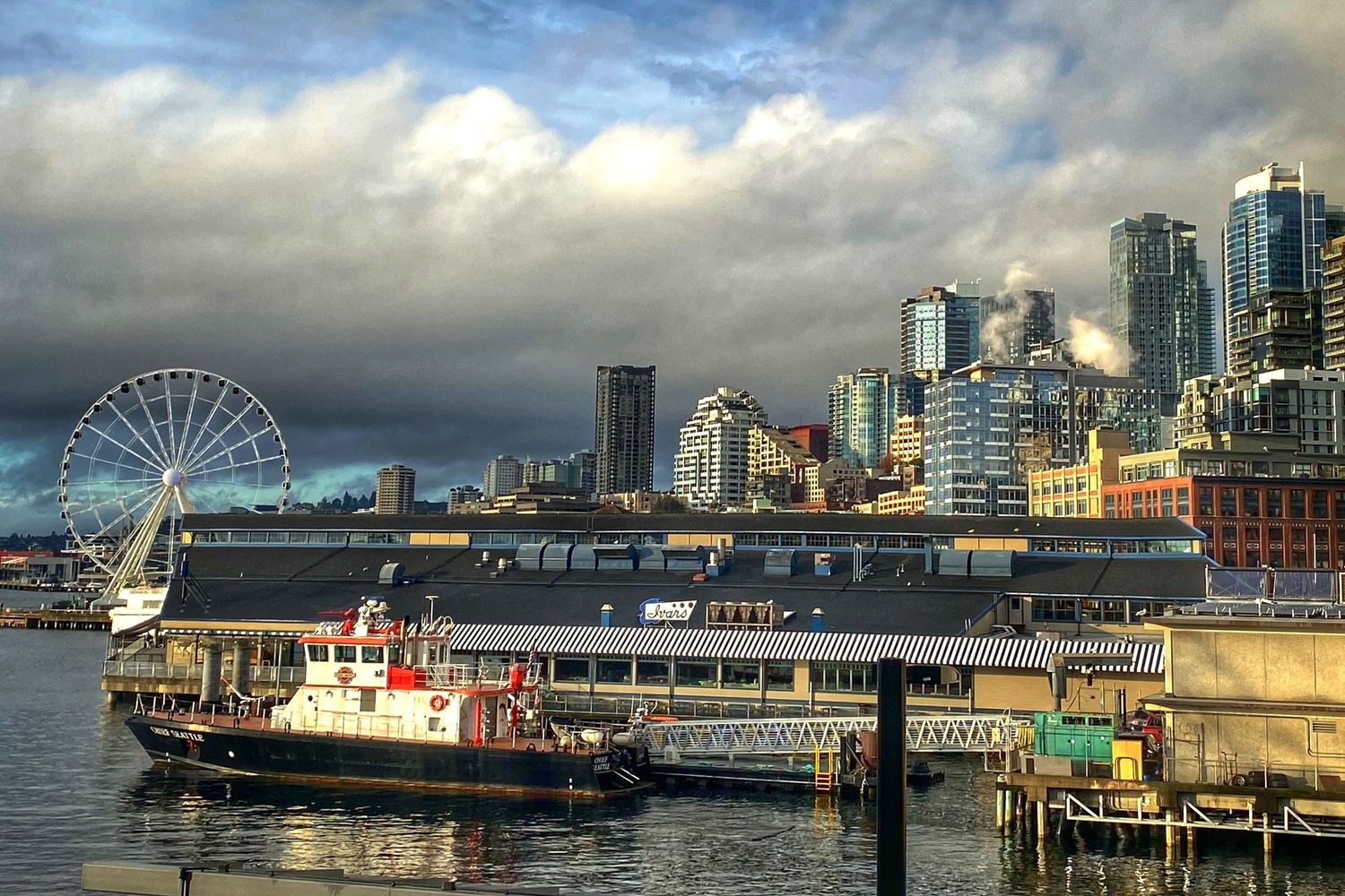 Panoramic view of Seattle's waterfront showing the Great Wheel, harbor boats, and downtown buildings under cloudy skies.