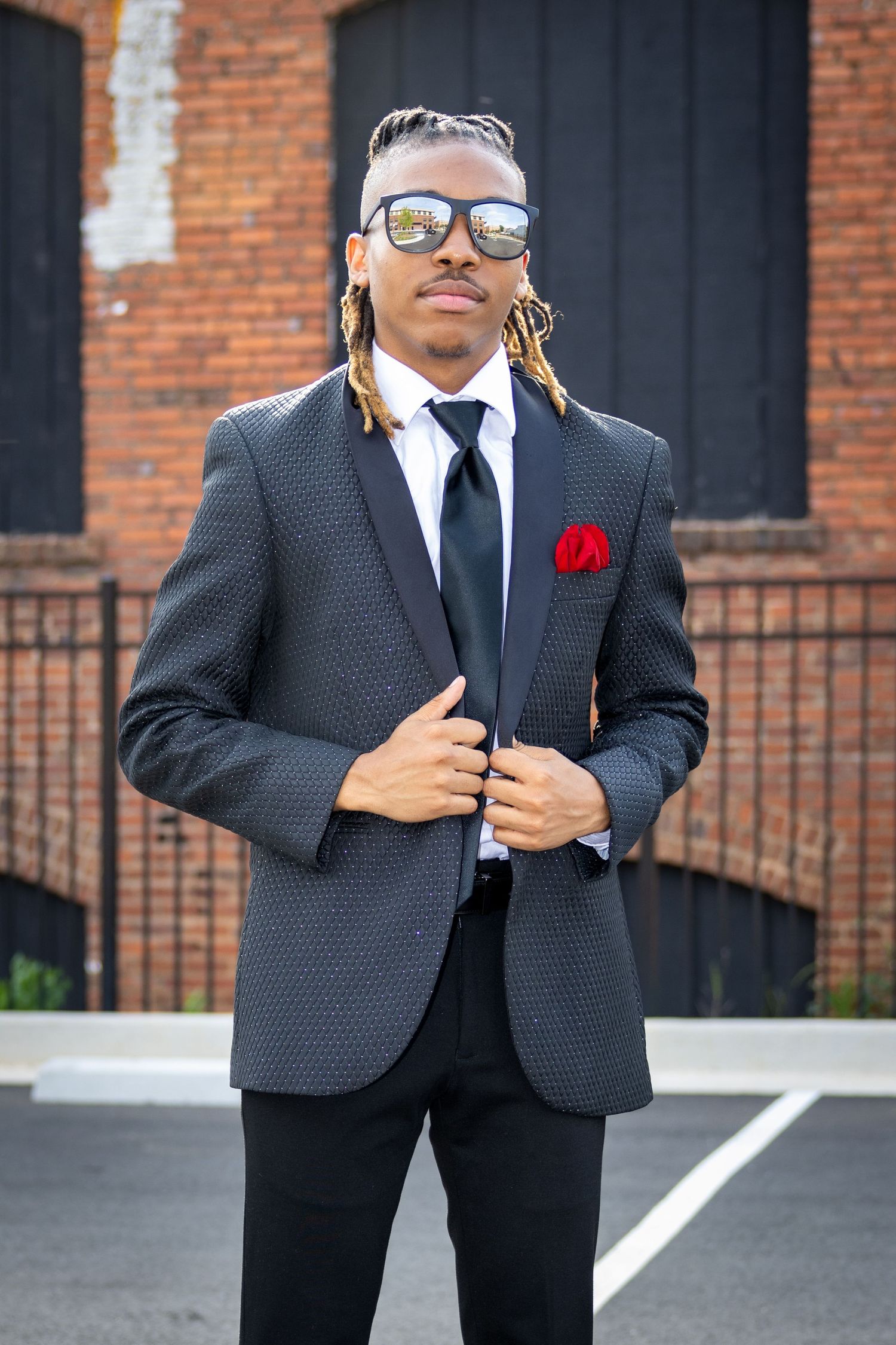 Stylish man in textured suit with sunglasses poses against brick wall.