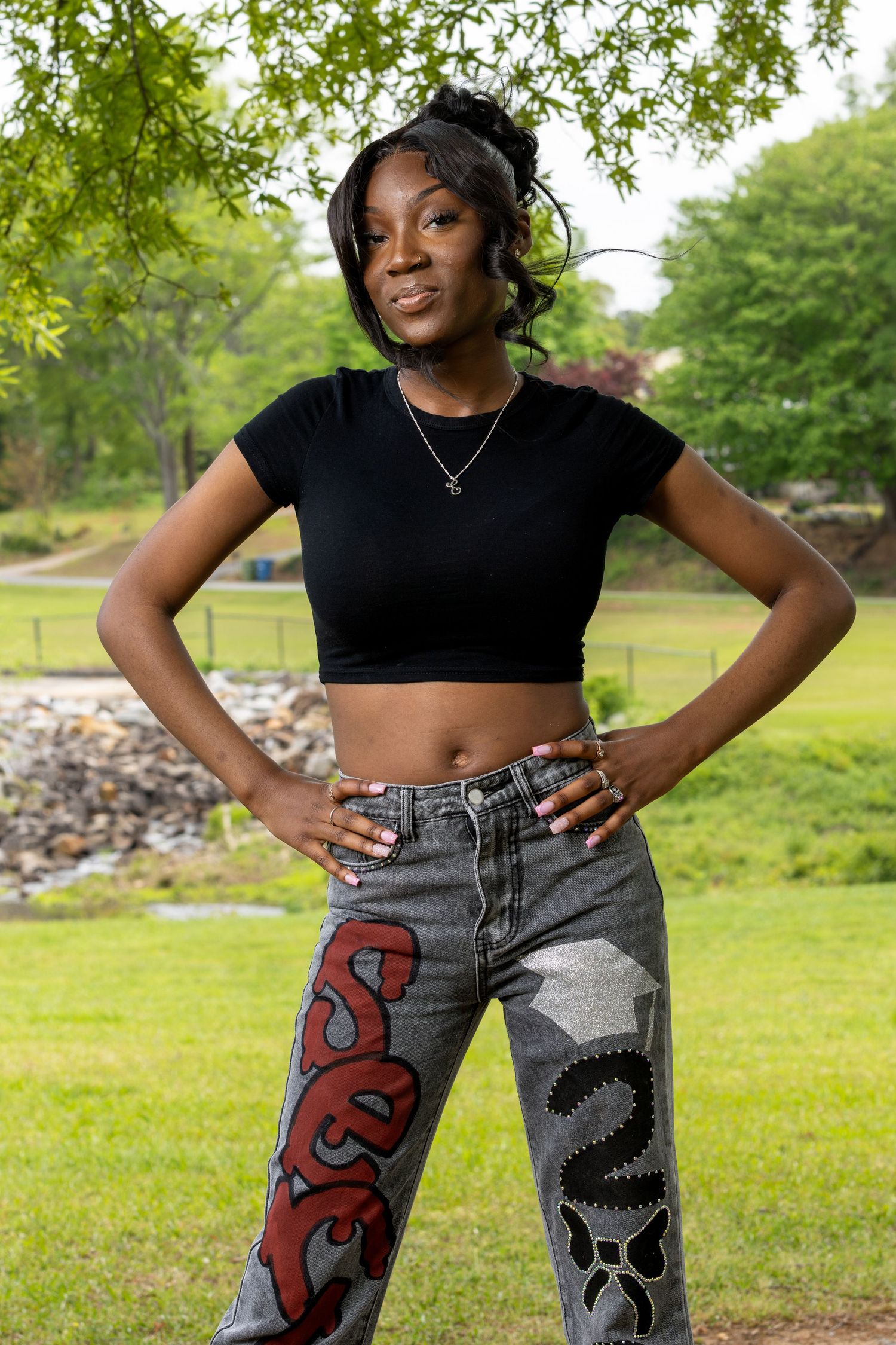 Model wearing black crop top and custom painted jeans poses in sunny park setting.