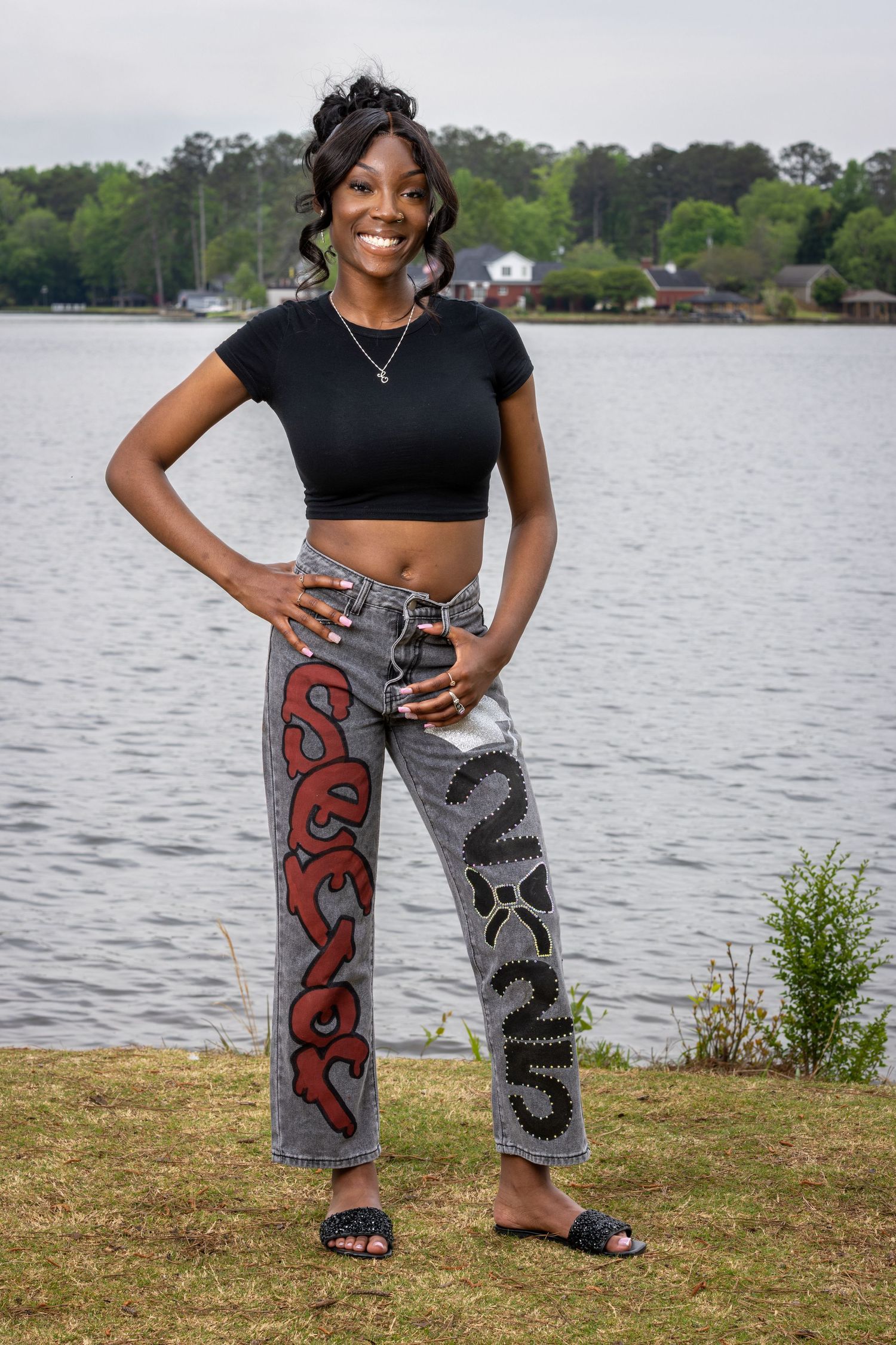Young woman posing by a lake in a black crop top and colorful snake-print jeans with slide sandals.