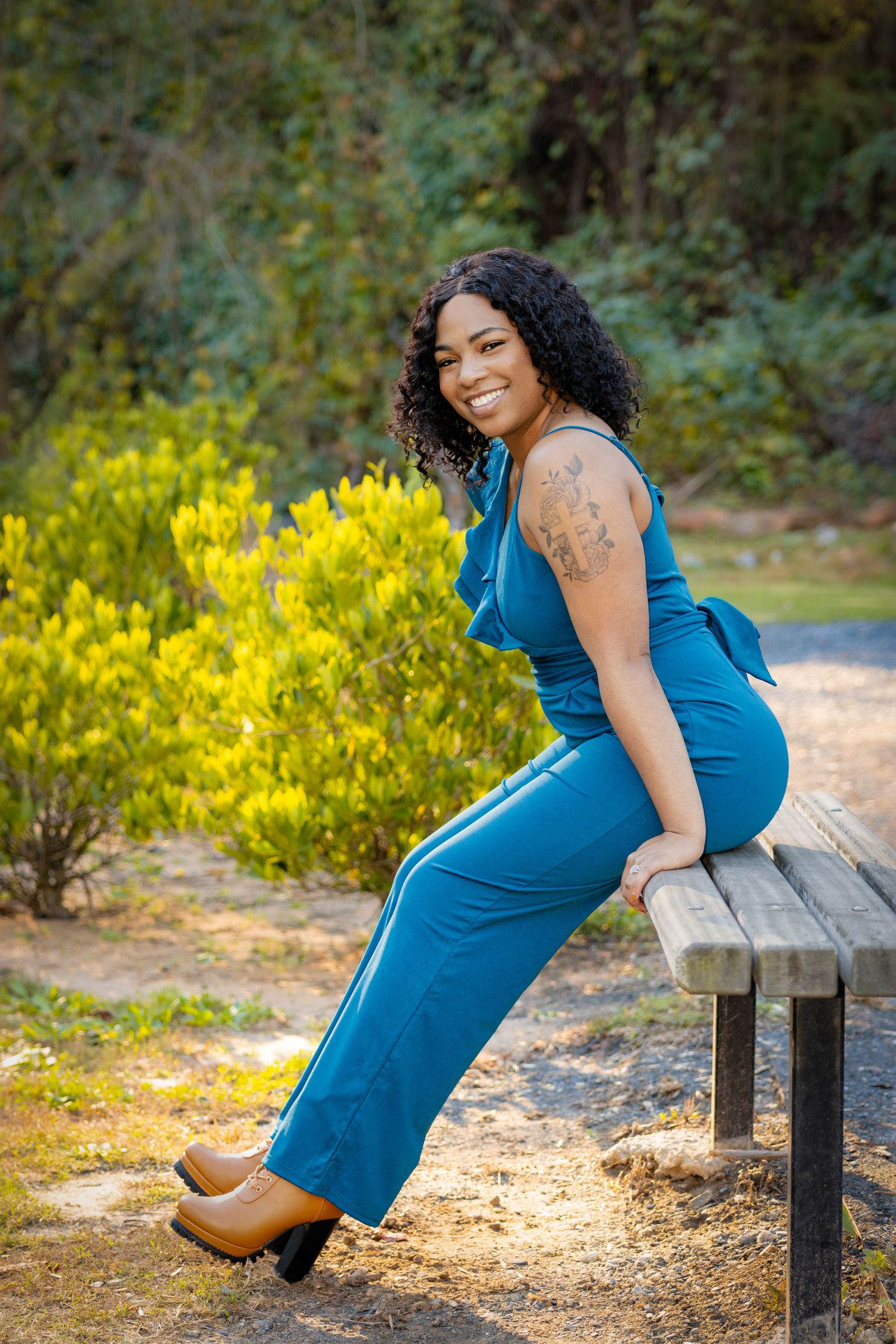 Woman in blue jumpsuit poses on wooden bench with yellow flowers in background.