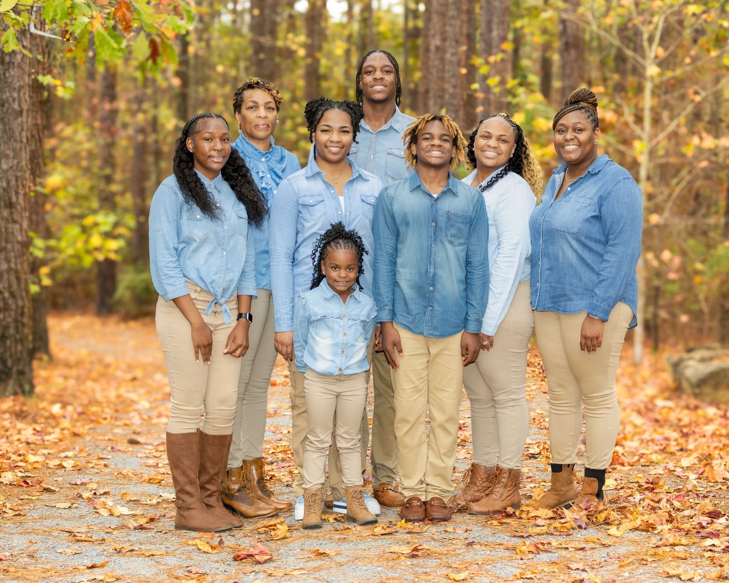 A group wearing coordinated denim and khaki outfits poses together in a wooded area with autumn leaves on the ground.