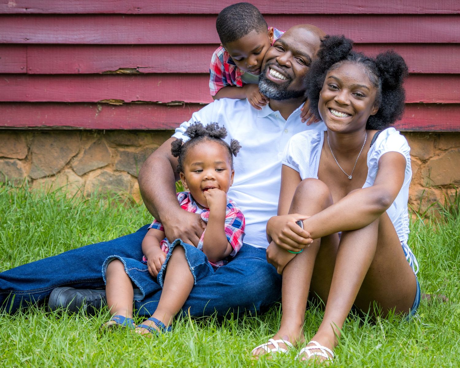 Family enjoying playful moments together while sitting on grass near a red barn wall.