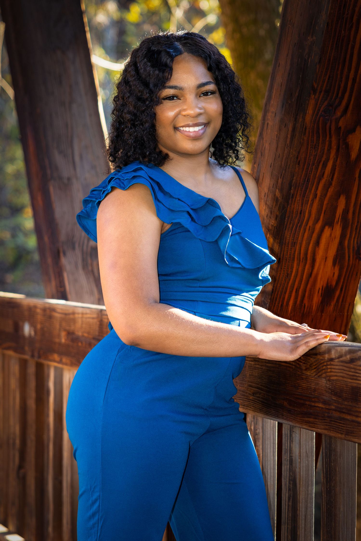 Woman in ruffled blue jumpsuit poses against wooden backdrop.