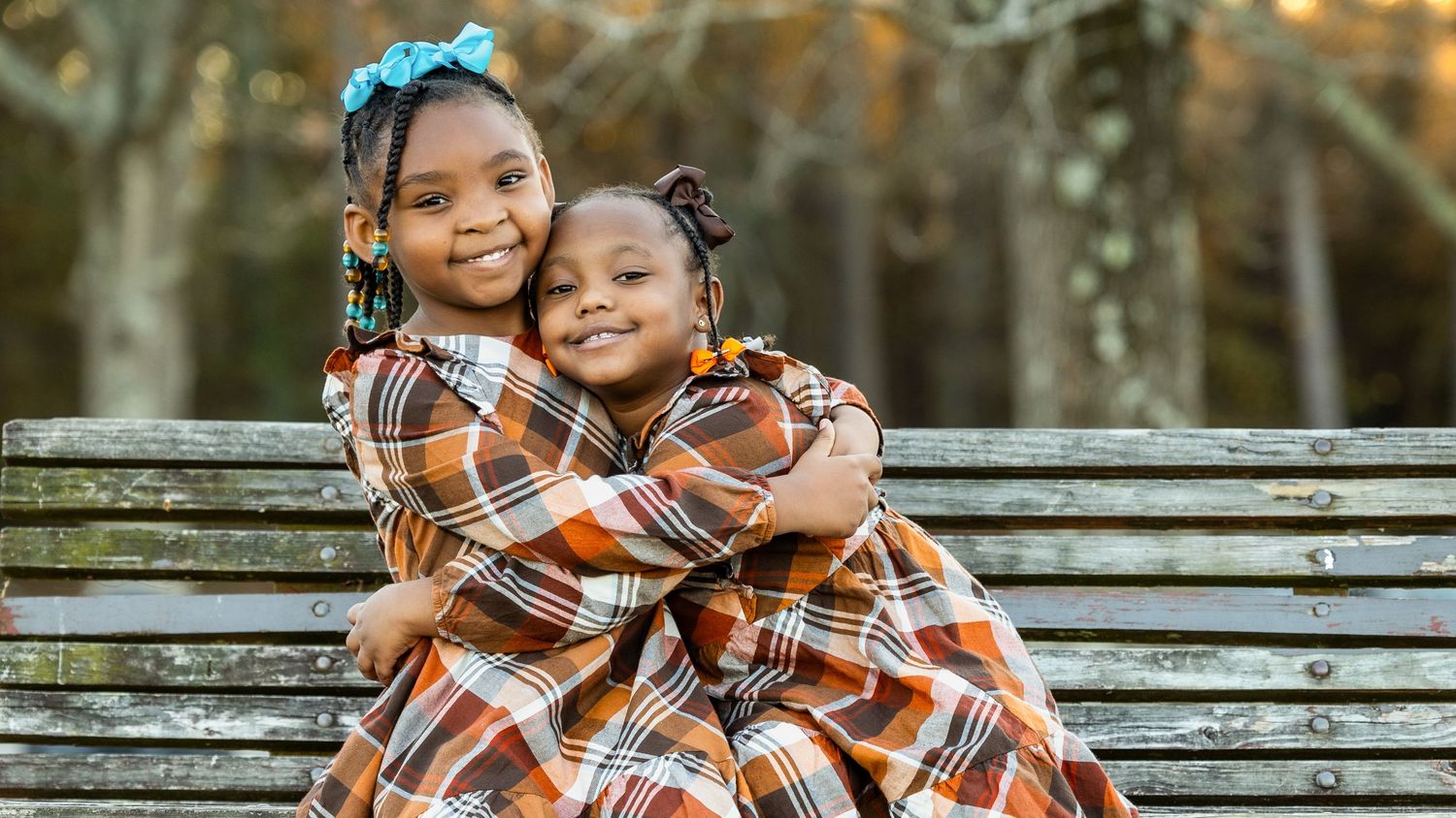 Two children in matching plaid outfits embrace on a wooden bench.