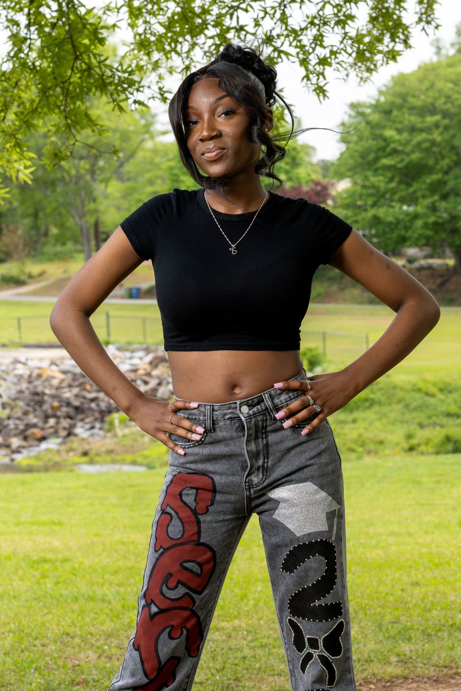 Young woman in black crop top and snake-print jeans poses confidently outdoors in a green park setting.