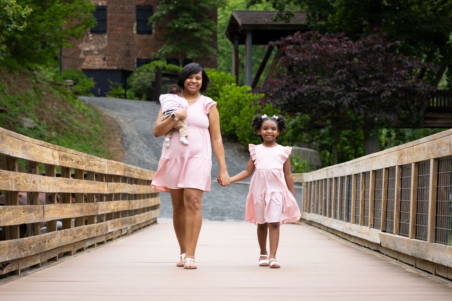 Mother and daughter walking hand in hand on wooden bridge wearing matching pink dresses.