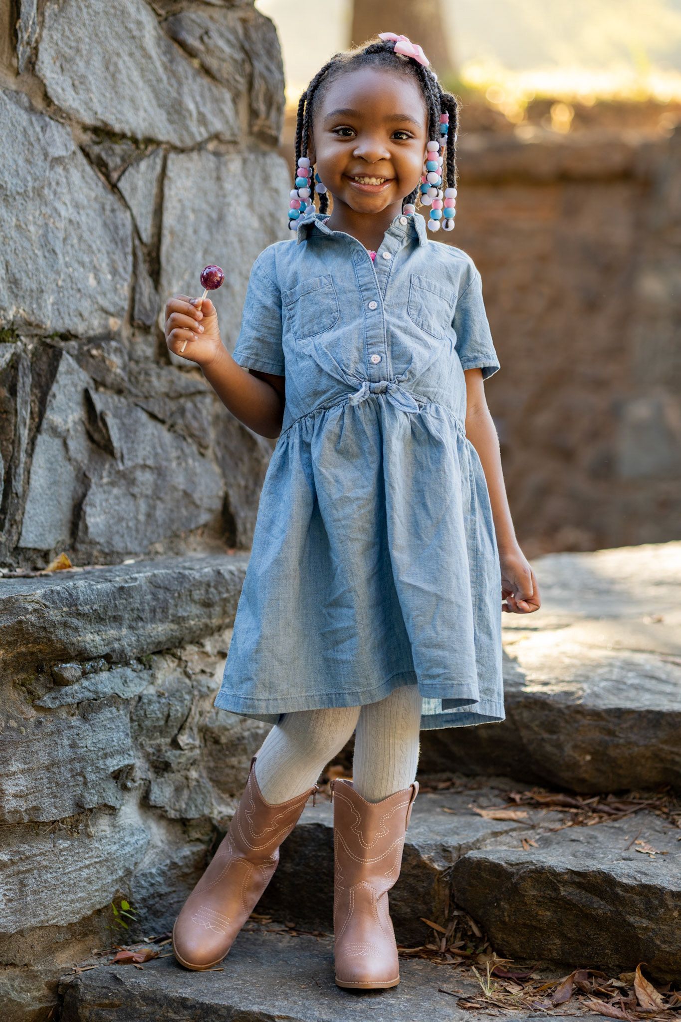 Small child wearing a light blue dress and white leggings poses near a stone wall at sunset.