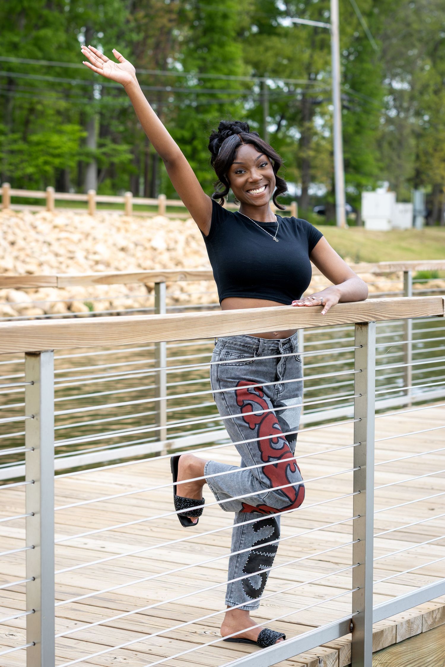 A smiling young Black woman poses on a wooden deck, wearing a black crop top and patterned jeans, with one arm raised.