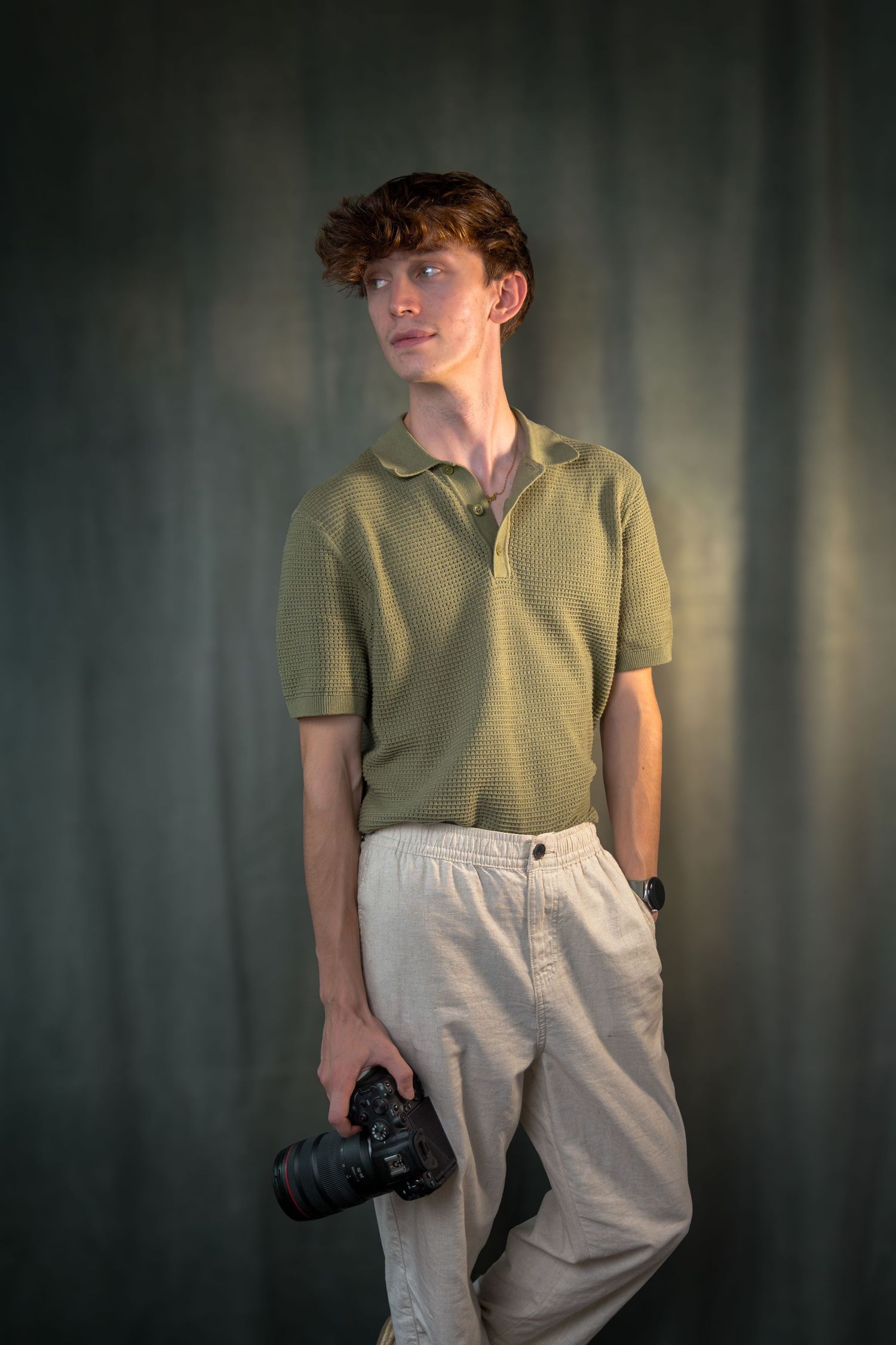 Person in olive shirt and light pants holds a camera in moody studio lighting.