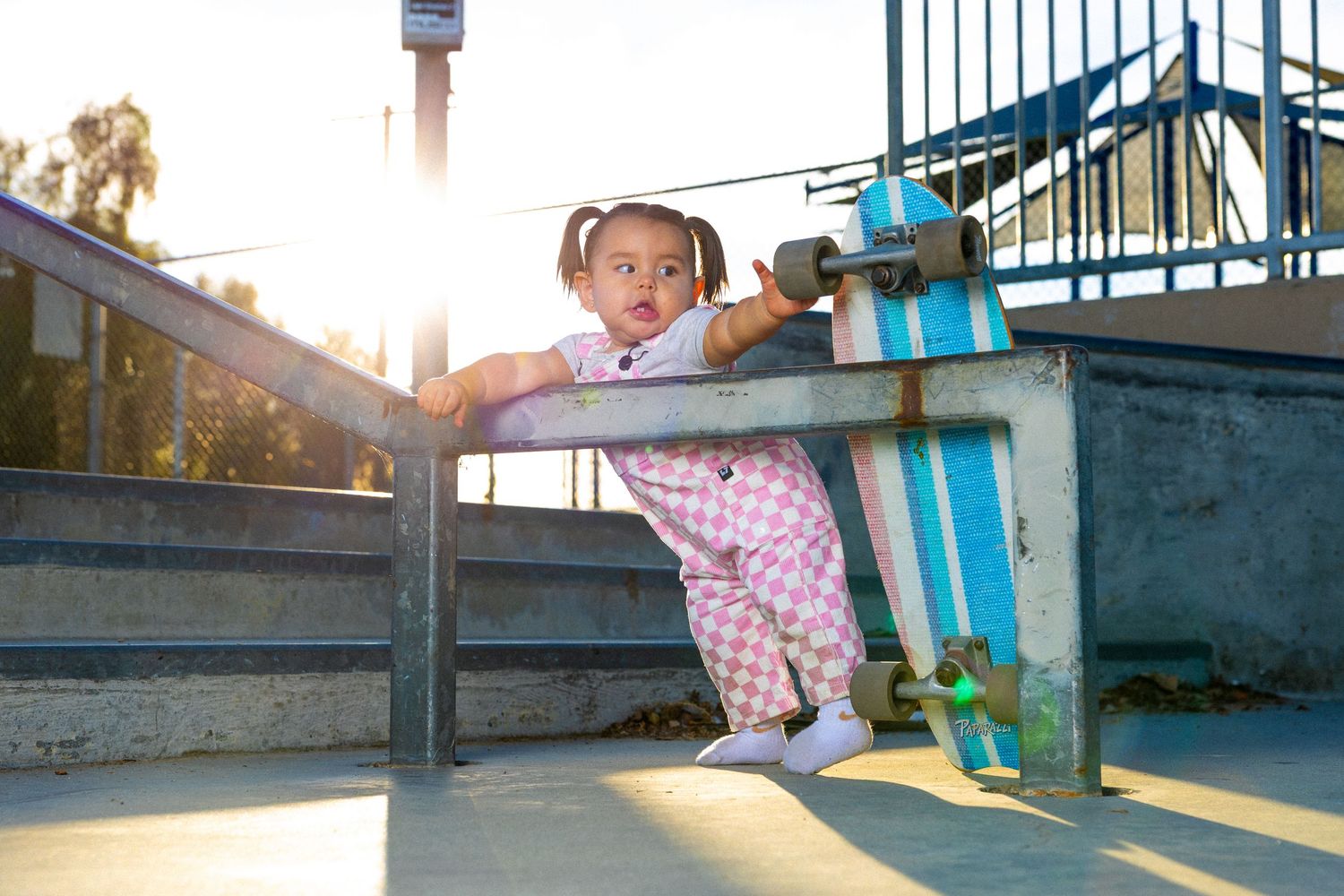 Baby girl leaning on a grinding rail in Moreno Valley Skate Park. She is reaching for the wheel of a skateboard that is leaning on the rail.