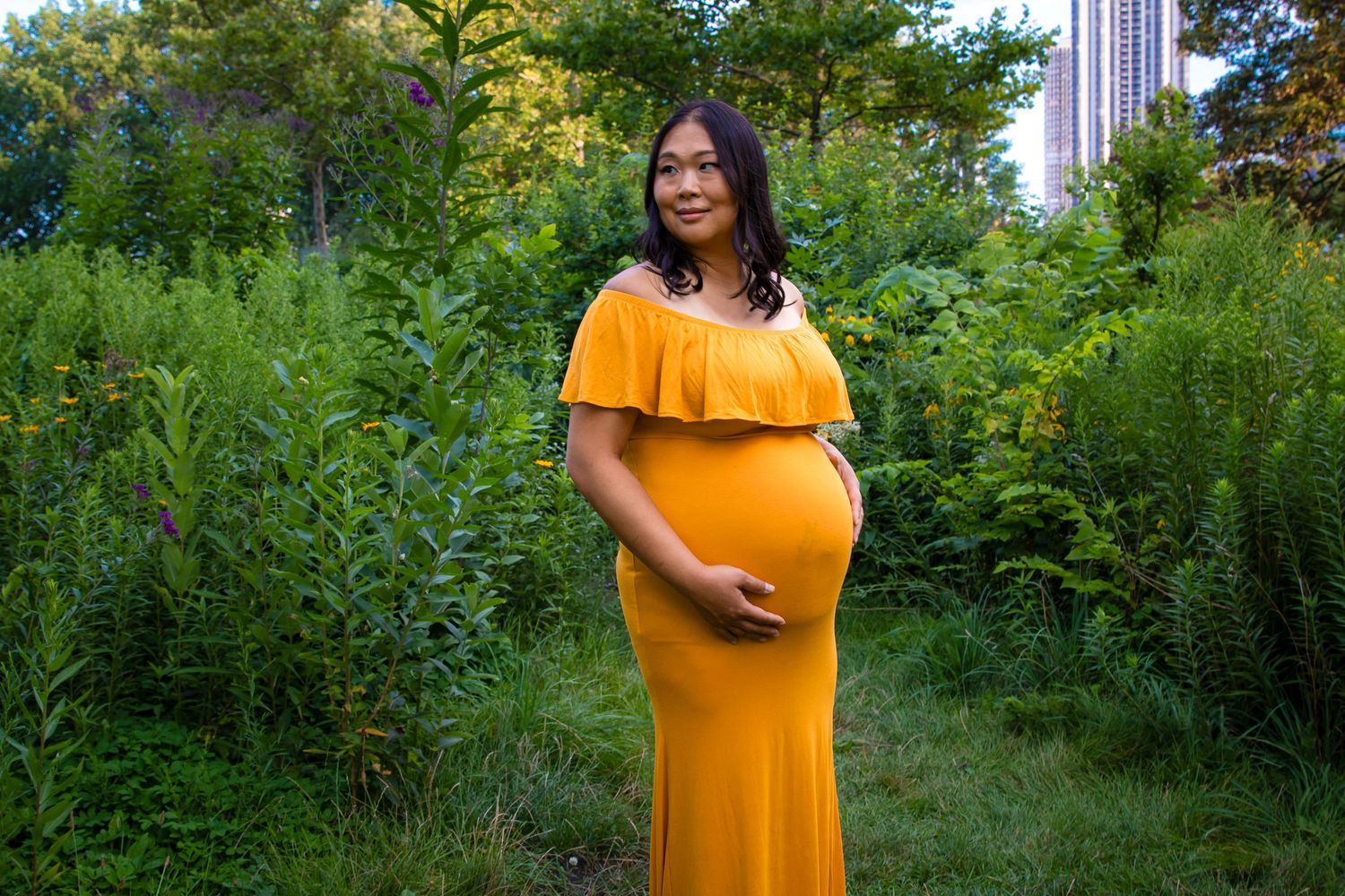 A pregnant woman in a yellow off-shoulder gown smiling while standing in a lush green garden.