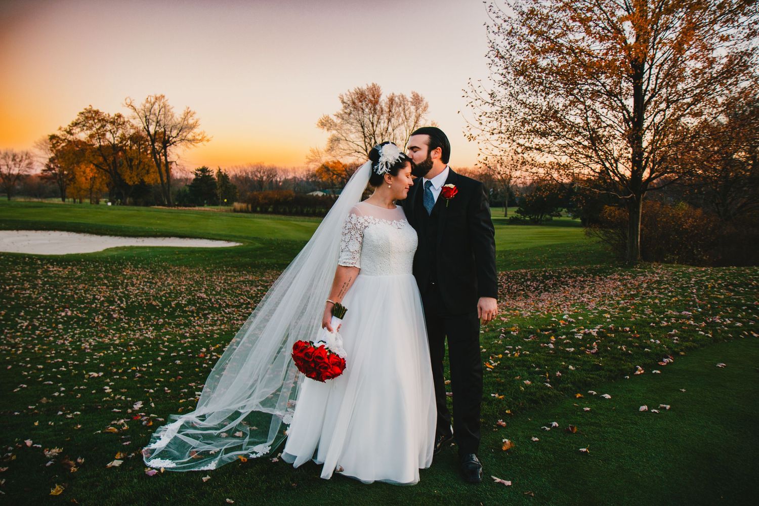 Bride and groom embrace romantically on golf course during vibrant orange and purple sunset.