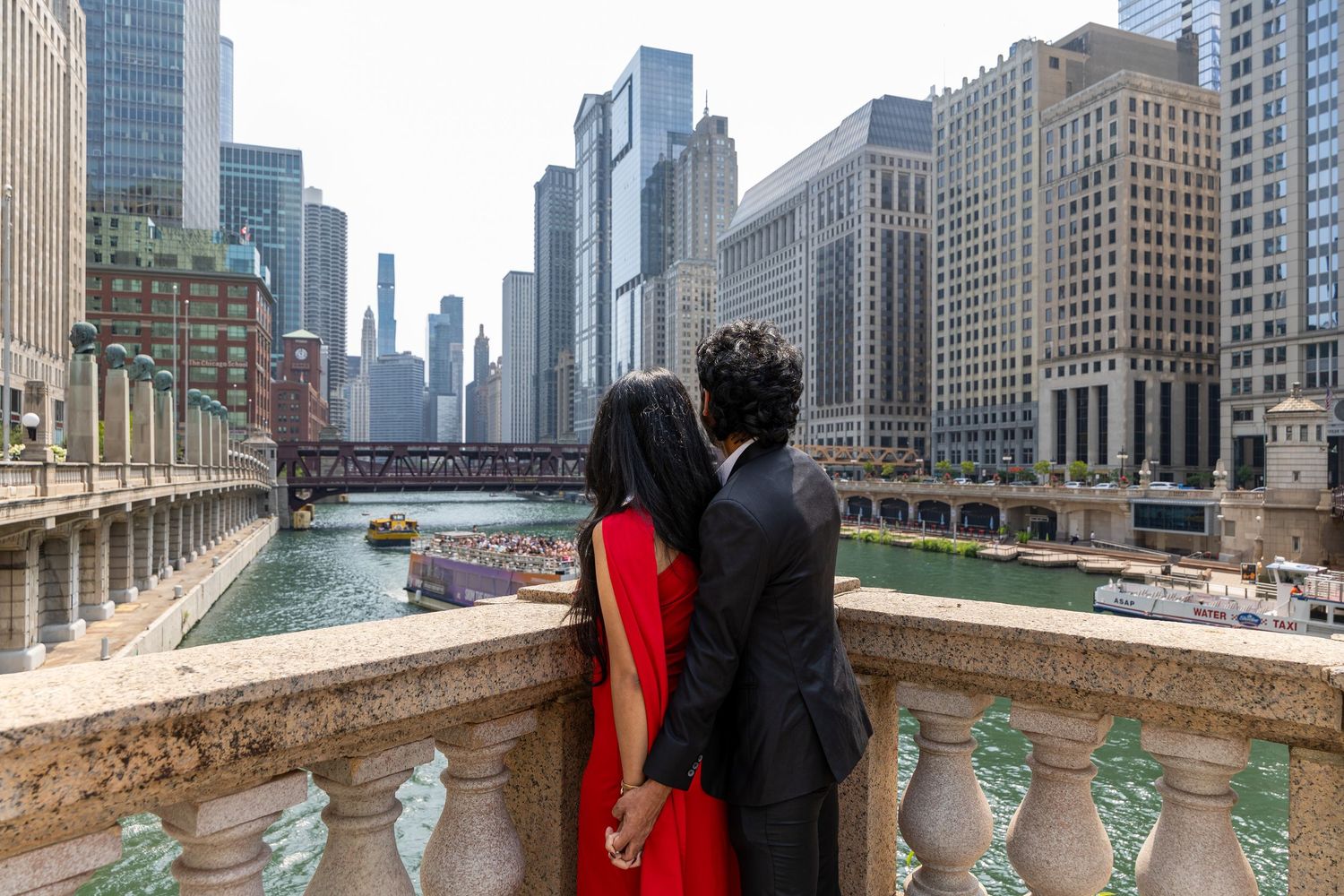 Couple holding hands overlooking Chicago River with city skyline, woman in red dress and man in black suit.
