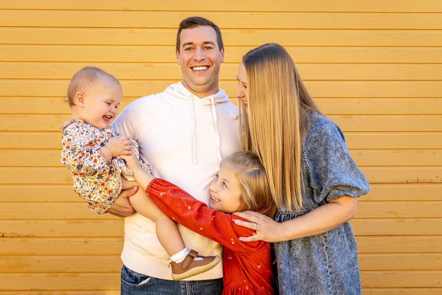 Joyful family of four posing together against a bright yellow wooden wall.