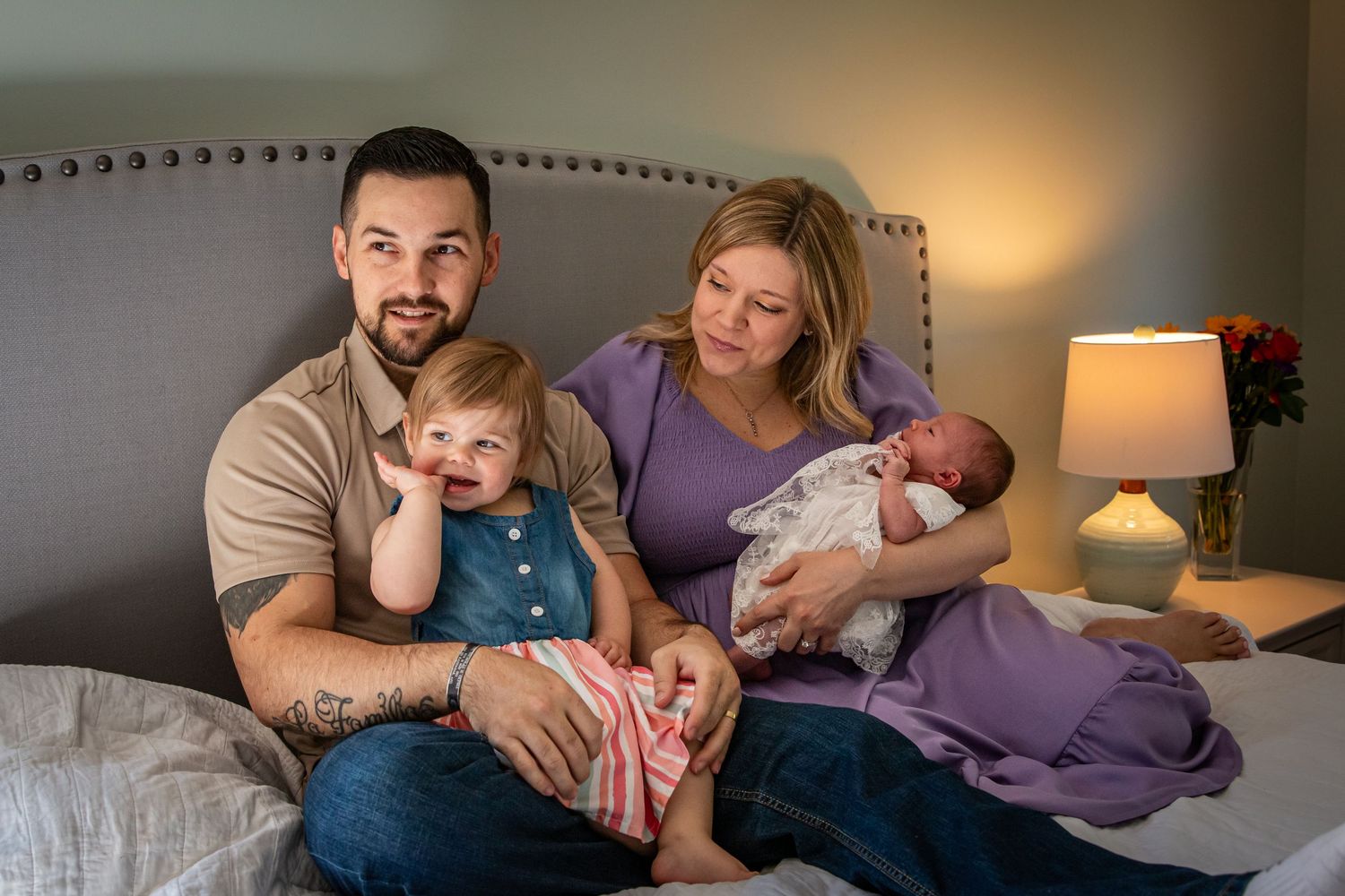 Young family sitting on a bed with a toddler and newborn baby in a warmly lit room.