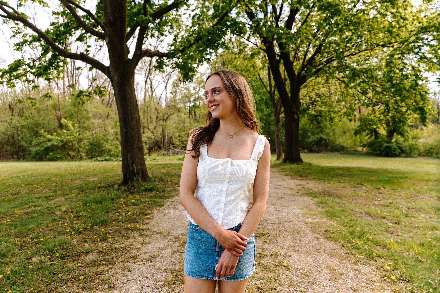 Young woman in white top and denim shorts smiling outdoors in a lush green park with large trees.