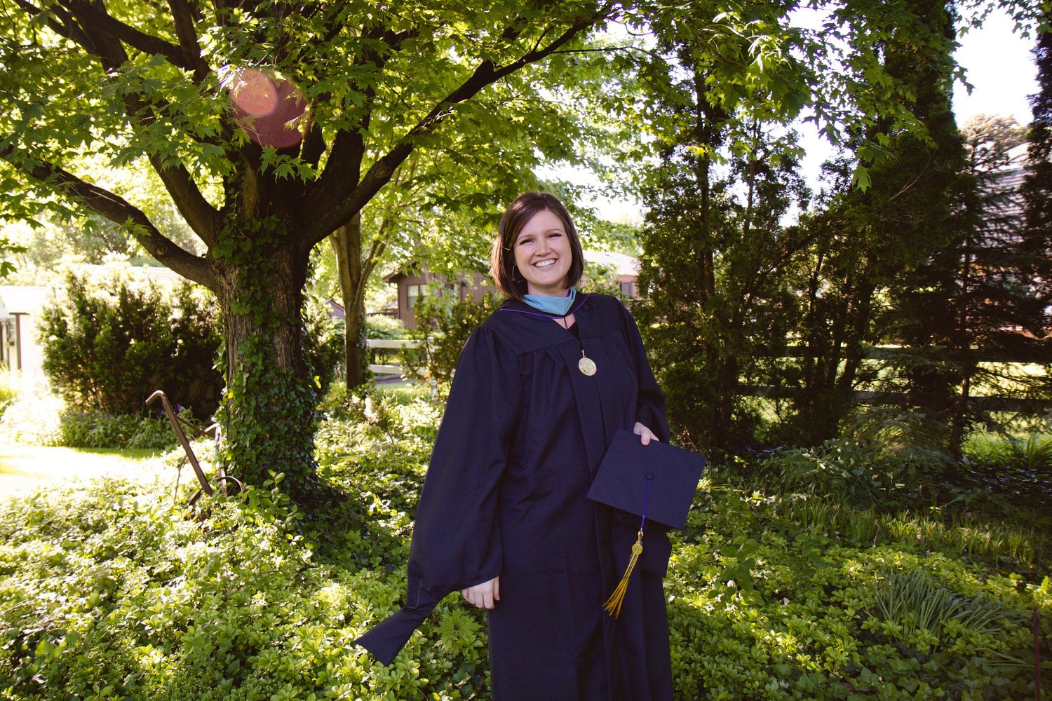 Woman in navy graduation gown and cap smiling outdoors among lush green trees and garden.