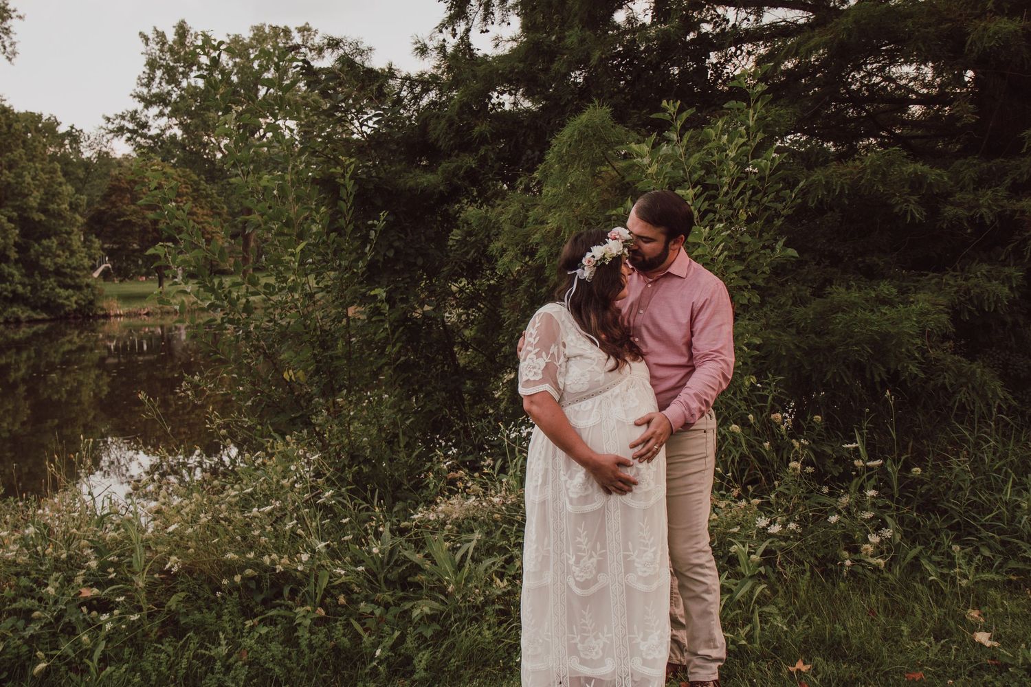Couple sharing a tender maternity moment by a pond surrounded by lush green trees.