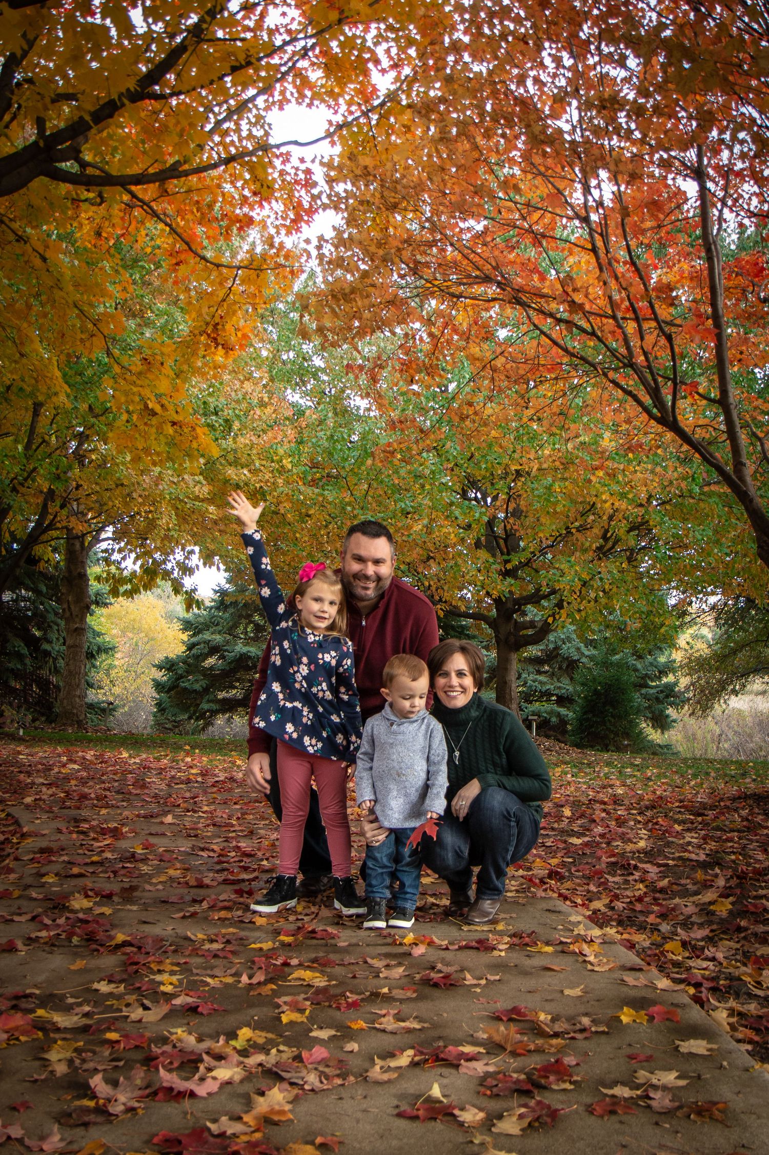 A happy family of four posing among vibrant autumn foliage with colorful fallen leaves on the ground.