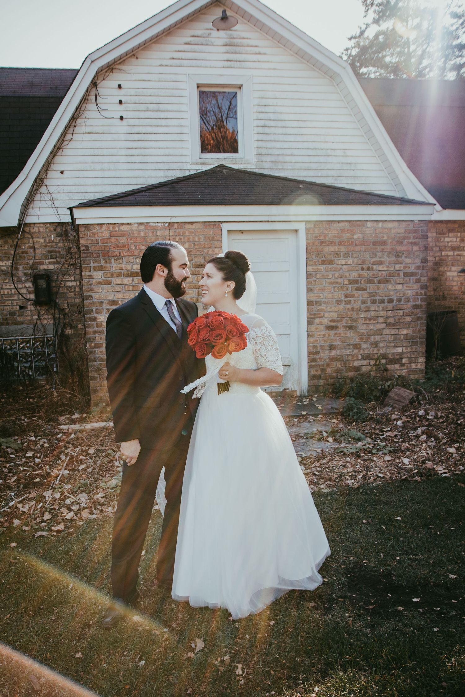 Bride and groom smile at each other holding red bouquet outside rustic brick barn in sunlight.