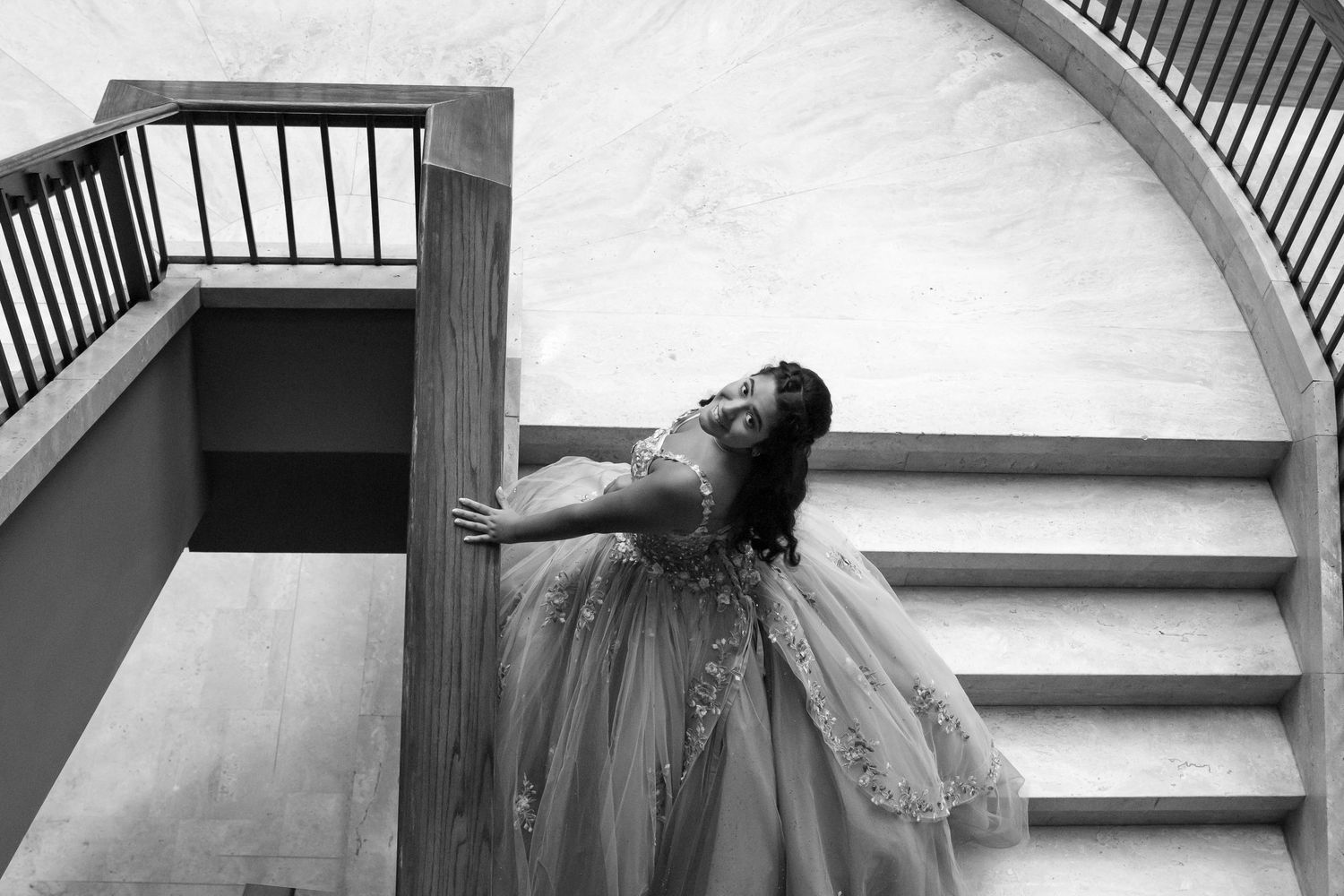 Black and white photo of a young woman in a ball gown posing gracefully on a spiral staircase.