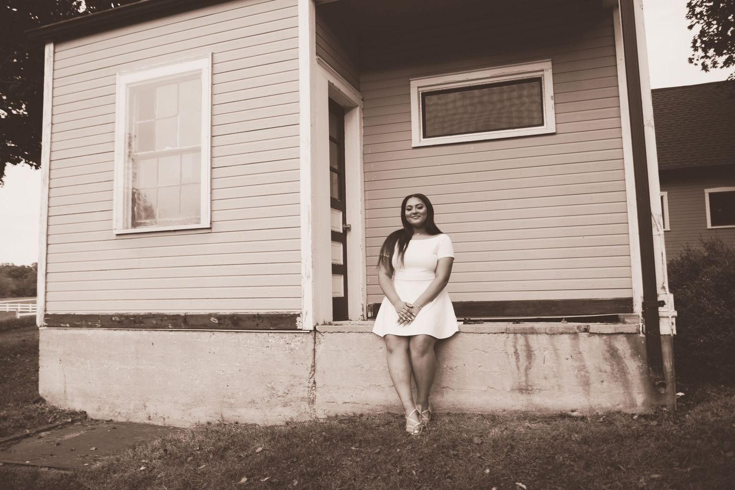 Young woman in white dress posing beside a charming vintage house exterior in a sepia-toned portrait photo.