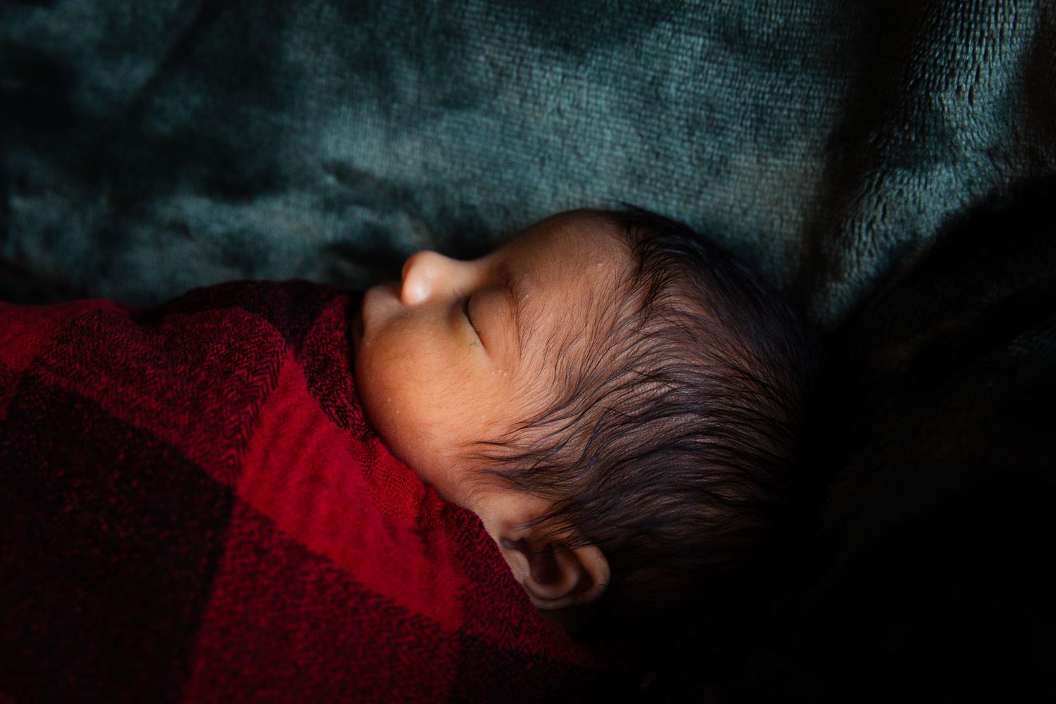 Peaceful newborn baby sleeping soundly wrapped in a red blanket on teal background.