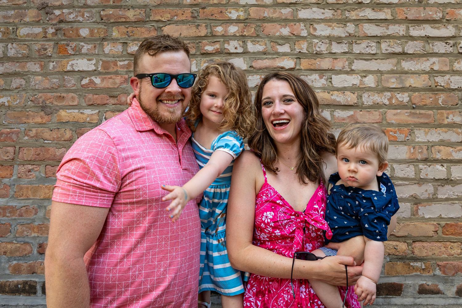 Happy family of four posing against a brick wall in colorful summer outfits.