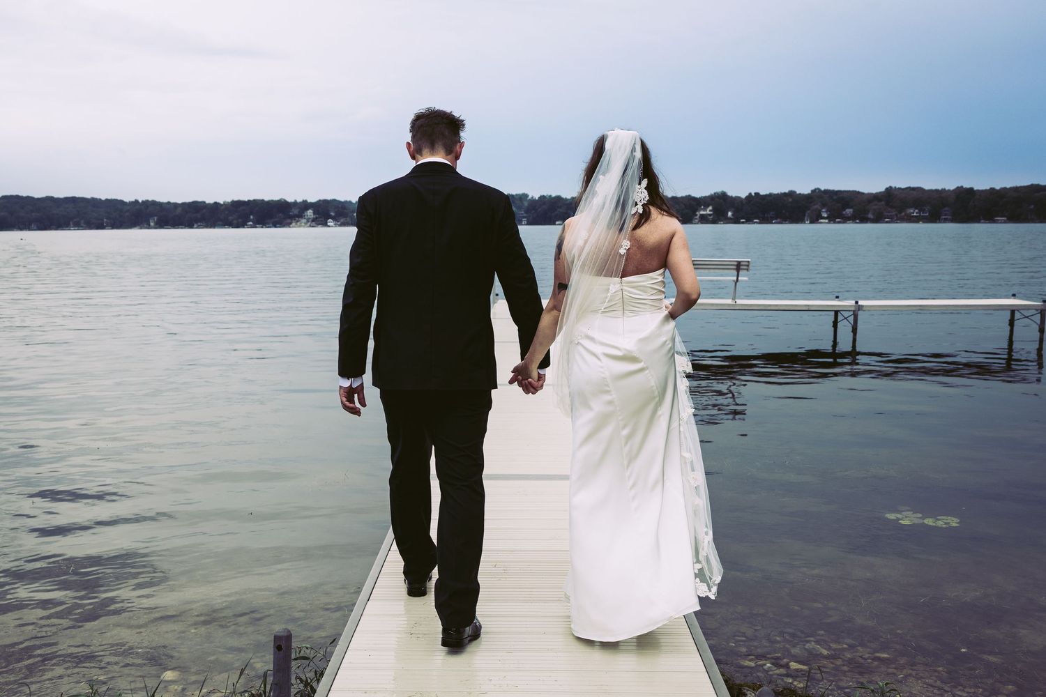 Bride in white gown and groom in black suit walk hand-in-hand on a lakeside dock together.