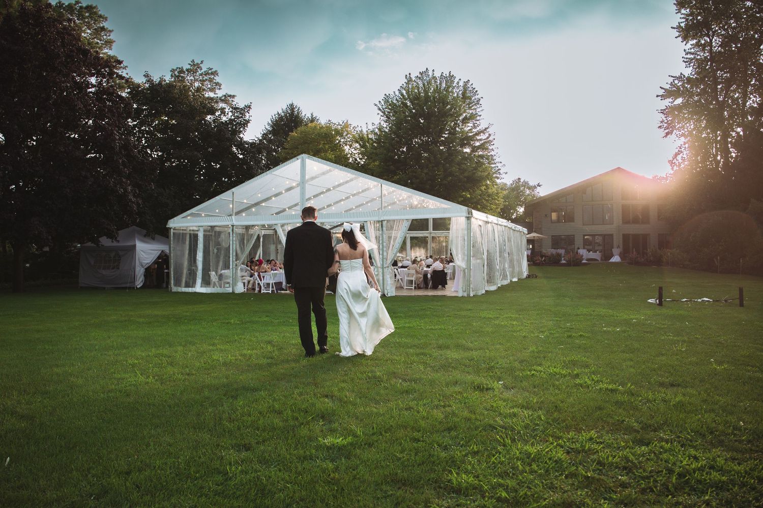 A bride and groom walk hand in hand toward a large white reception tent glowing in golden sunset light.