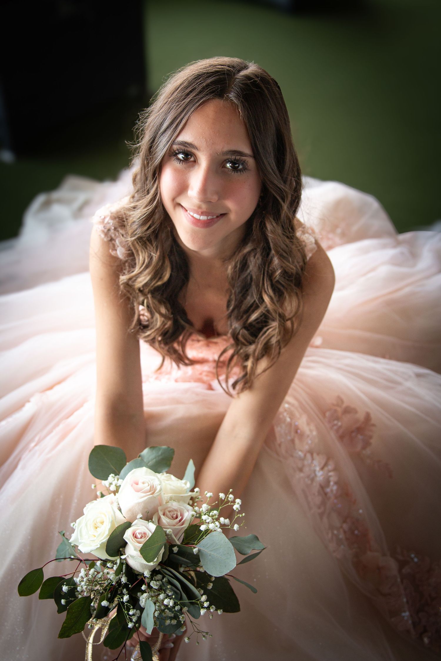 Smiling young woman in blush pink quinceañera ball gown holds white and pink rose bouquet while seated gracefully.
