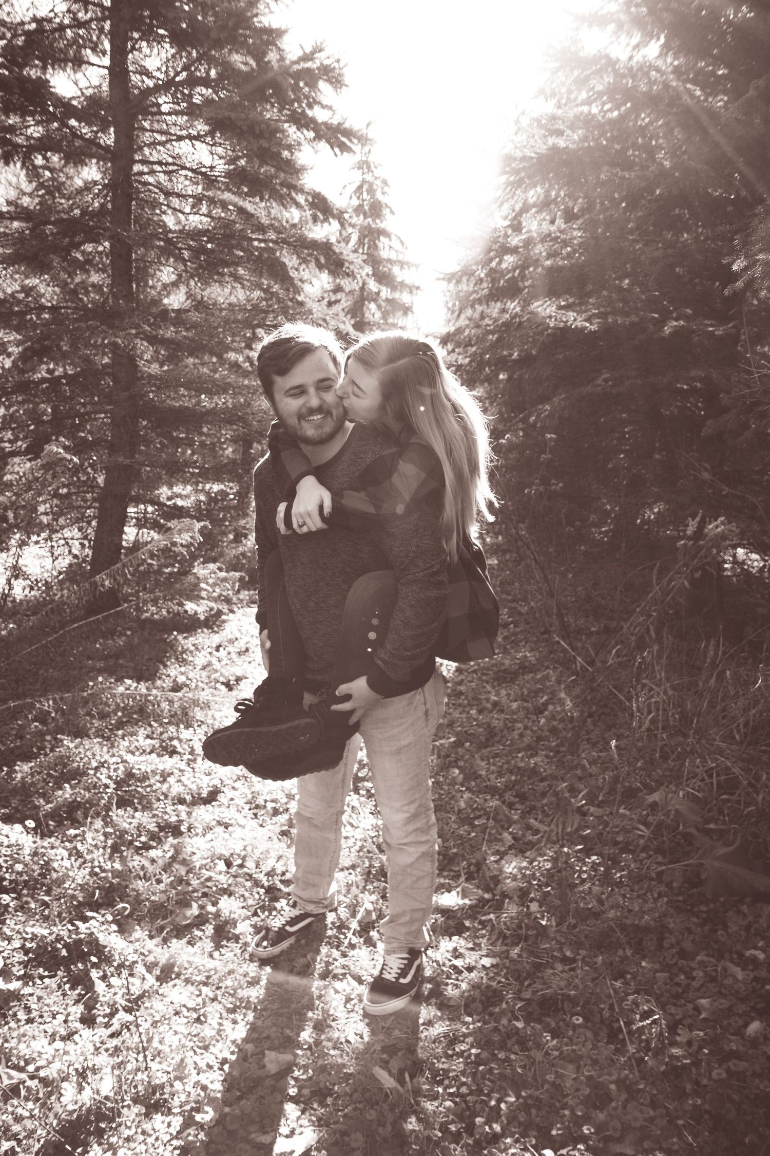 Black and white photo of a couple in a forest, woman kissing man on cheek as he carries her piggyback.
