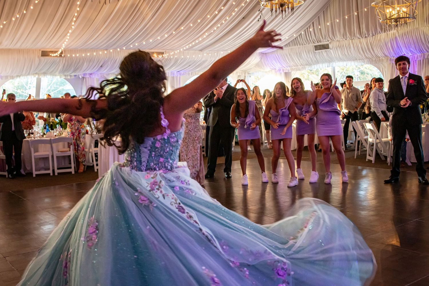 Quinceañera girl twirls in teal floral ball gown on dance floor surrounded by court in lavender dresses at reception.