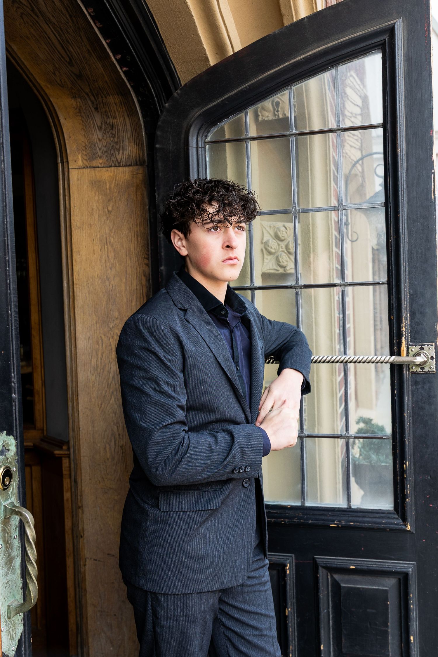 Young man in gray suit with curly hair leaning against ornate black door with crossed arms.