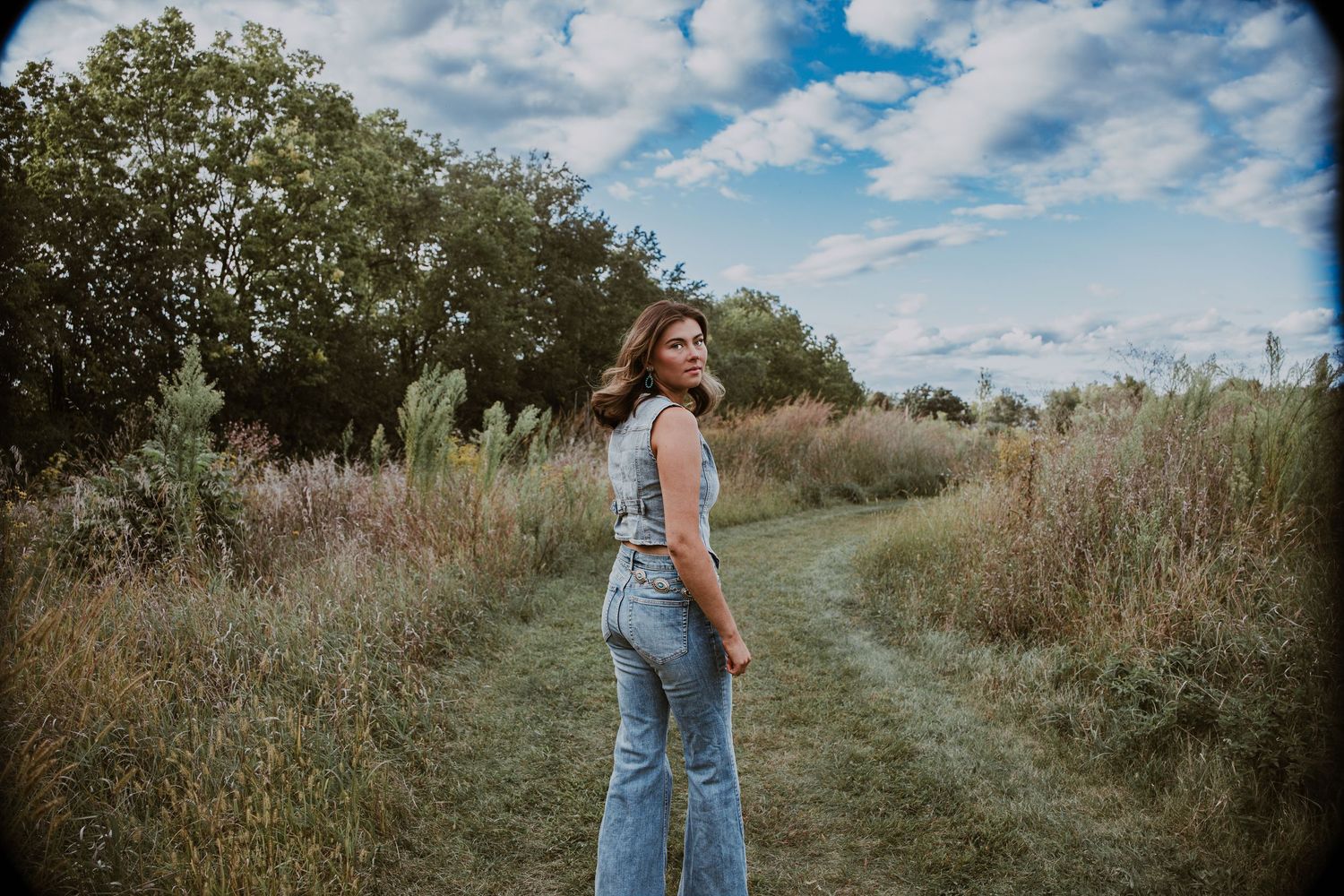 Young woman in flare jeans stands on a grassy field path, looking back over her shoulder under a cloudy sky.