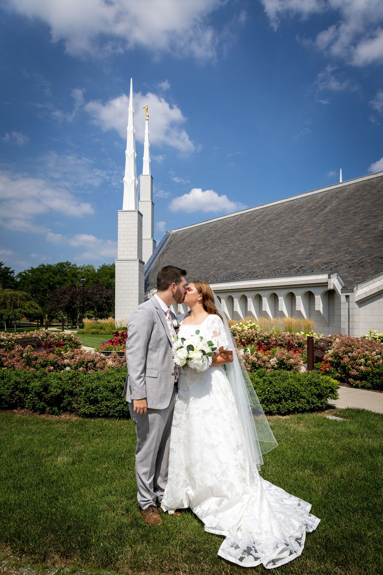A bride and groom share a kiss in front of a white LDS temple with tall spires and colorful gardens.