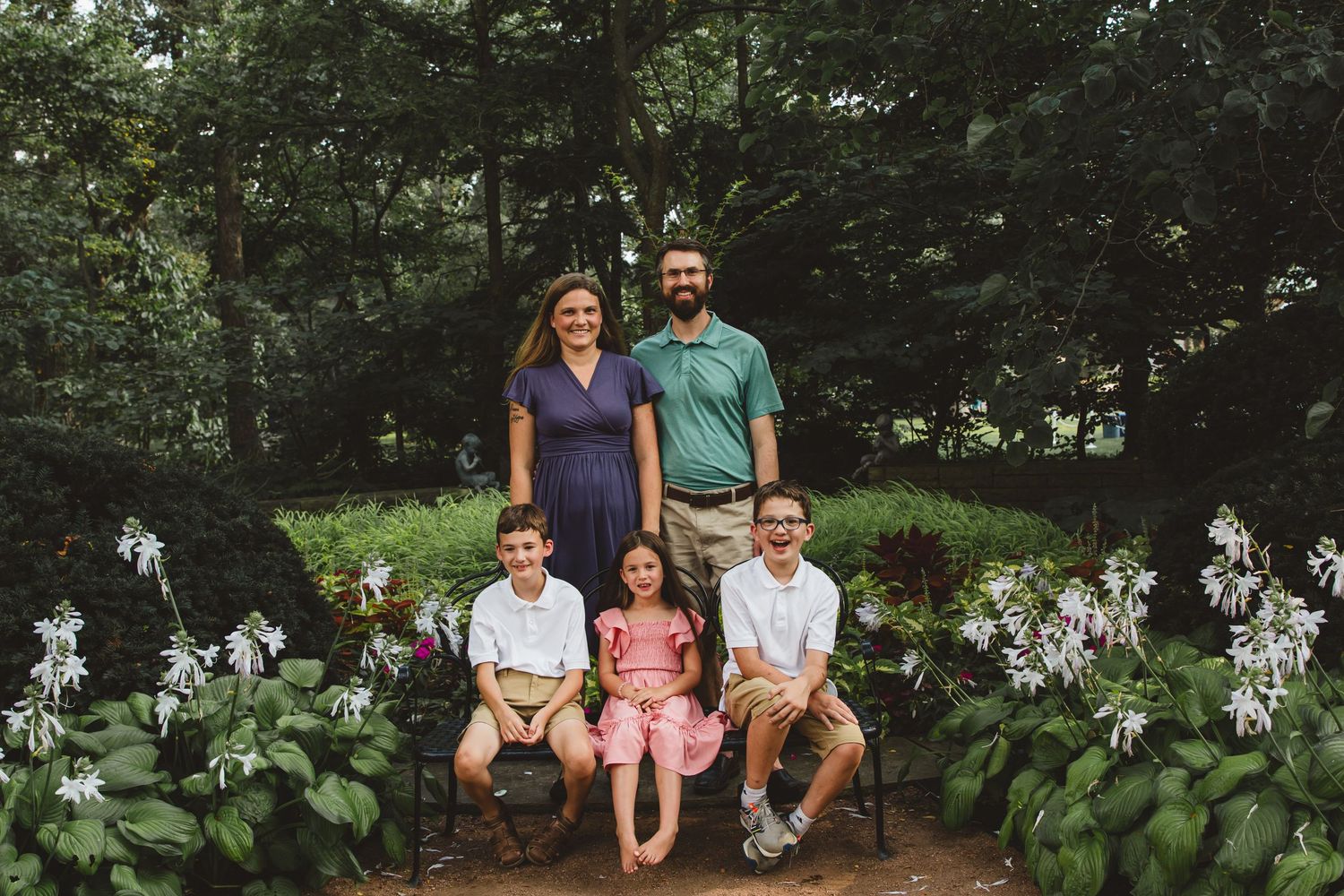 Family of five posing in a lush garden surrounded by white flowers, with three children seated on a bench.