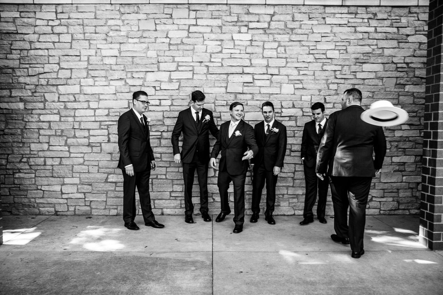 Black and white photo of groom and groomsmen in dark suits posing casually against brick wall.