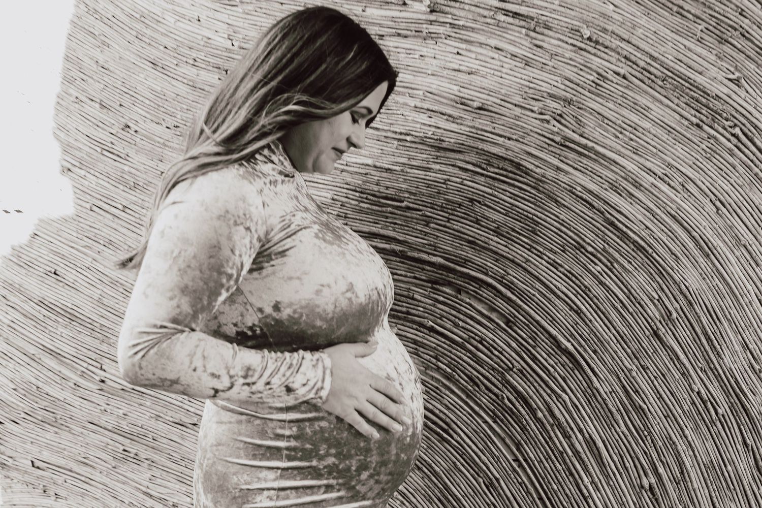 Artistic sepia maternity photo of pregnant woman in lace dress holding baby bump against textured background.