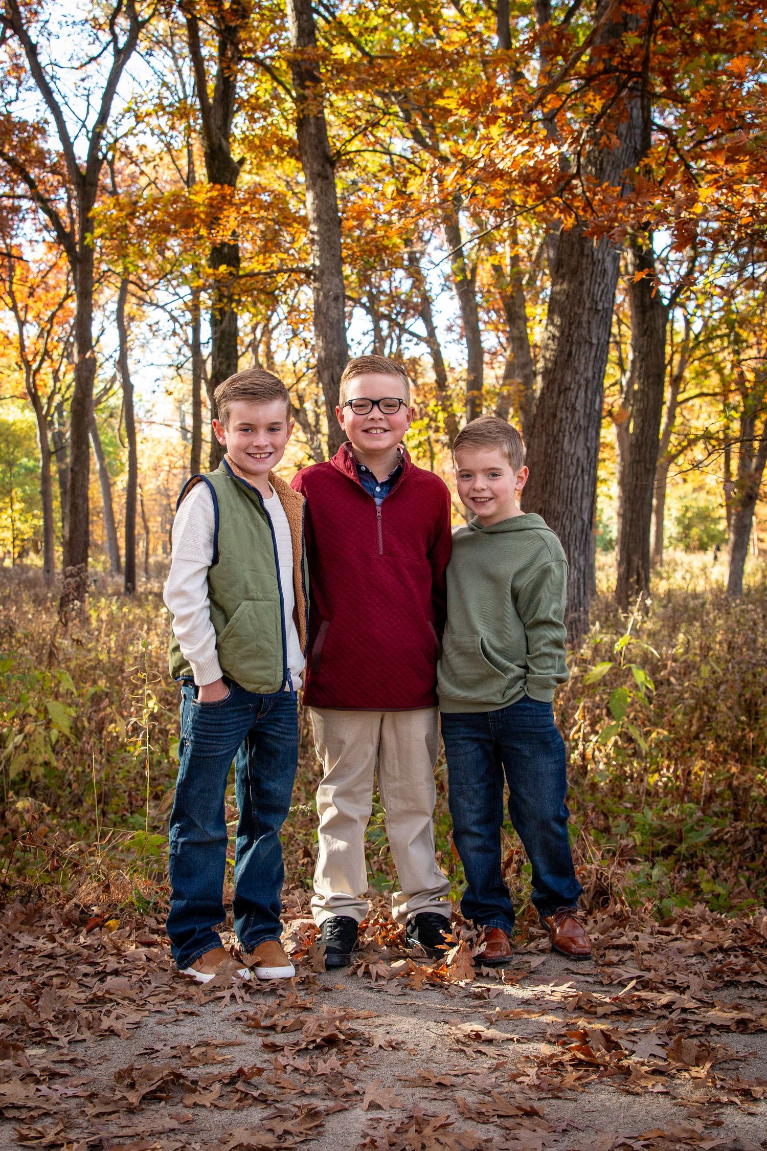 Three brothers smiling together on a leaf-covered path in autumn woods.